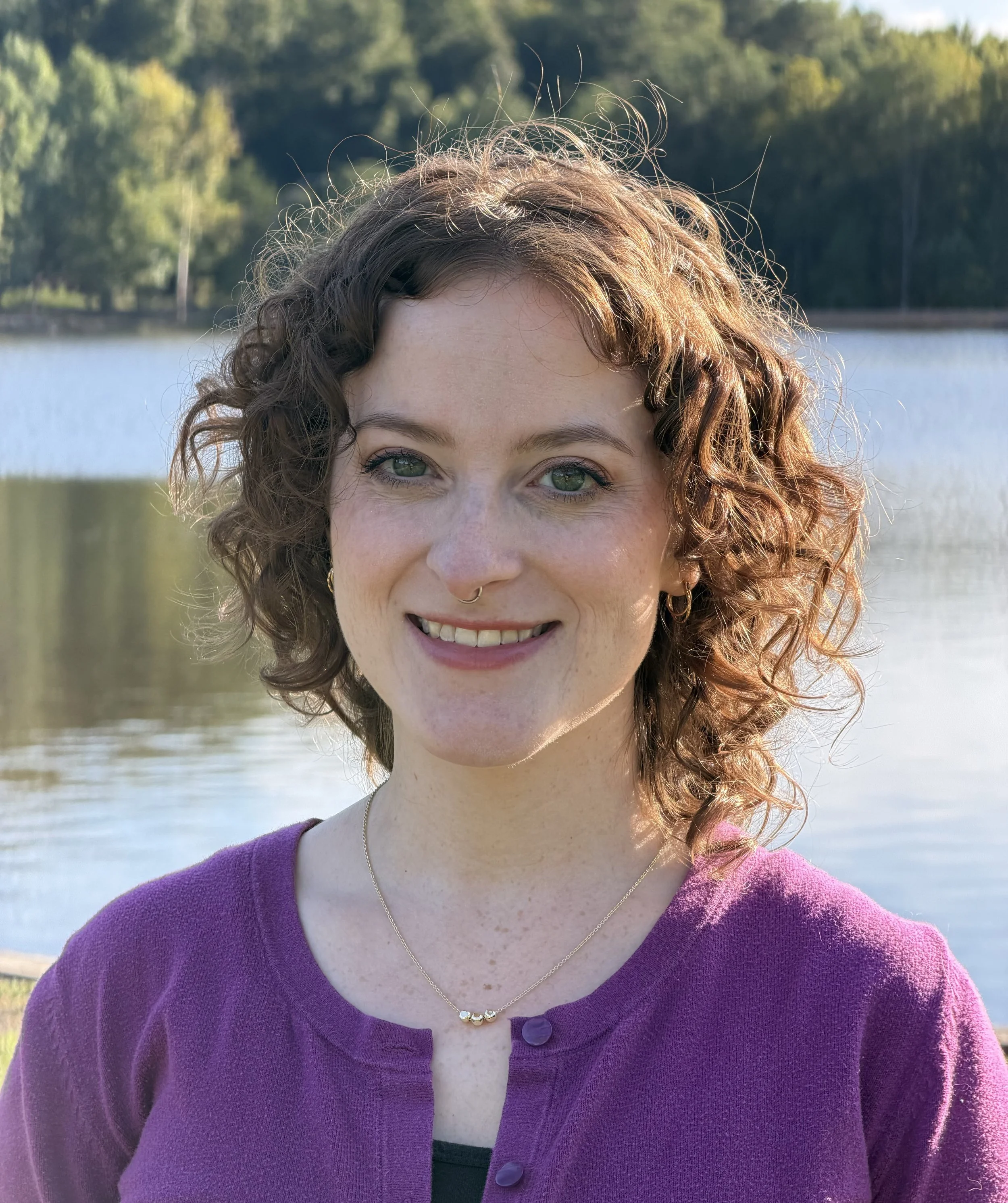 A woman with curly red hair and blue eyes is smiling outdoors near a body of water, wearing a purple cardigan and a necklace with three beads.