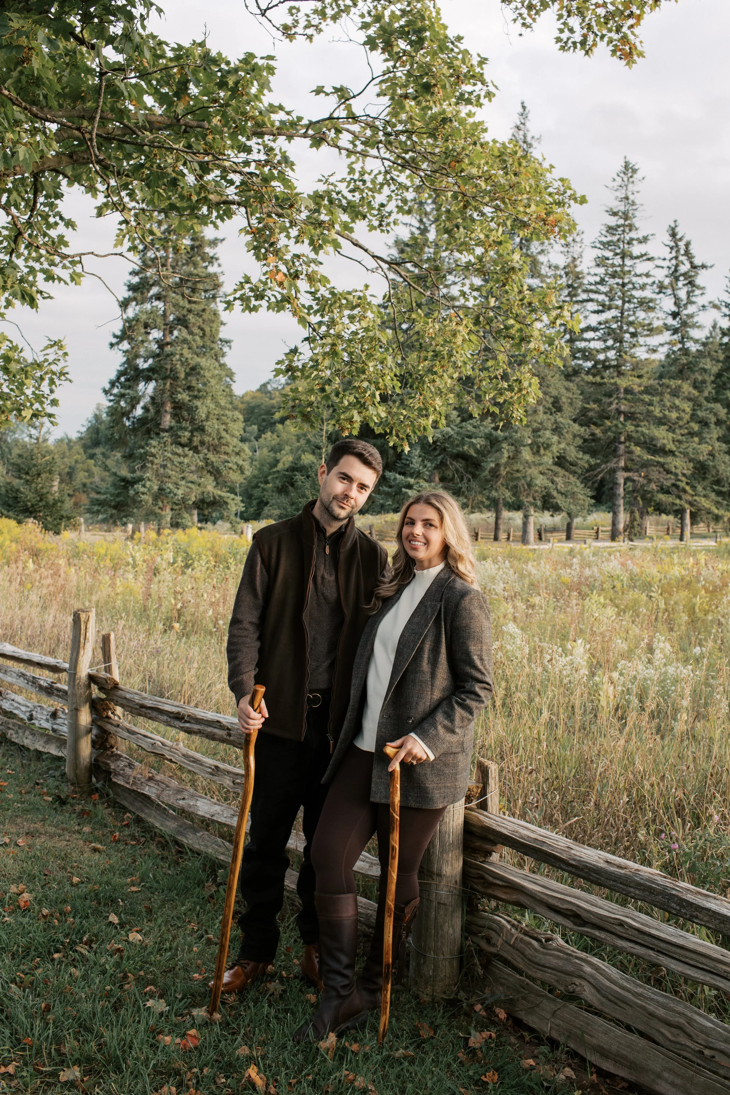 Husband and wife, Joel and Brittany, standing outdoors in a grassy area with trees in the background, holding wooden canes and smiling.