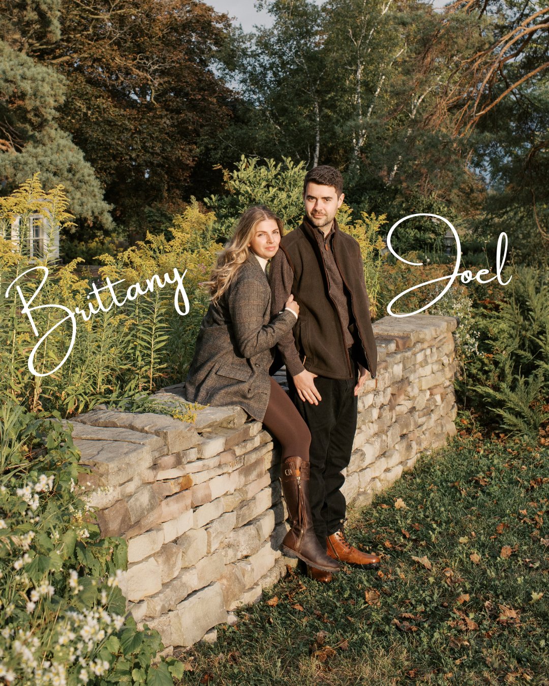 Husband and wife, Joel and Brittany, in fall clothing stand and sit on a stone wall in a garden with trees and bushes.