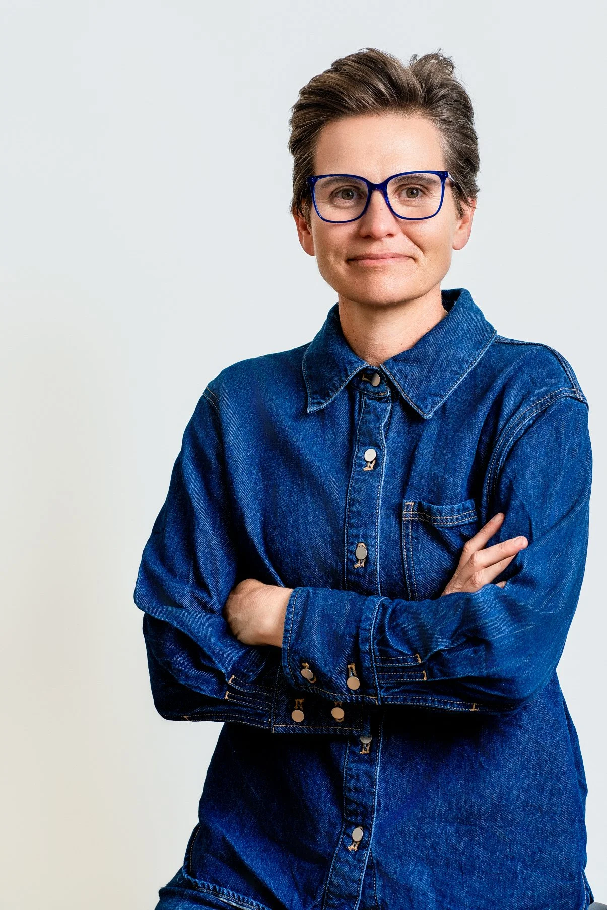 A woman with short hair and blue glasses wearing a denim jacket, posing with arms crossed against a plain light background.