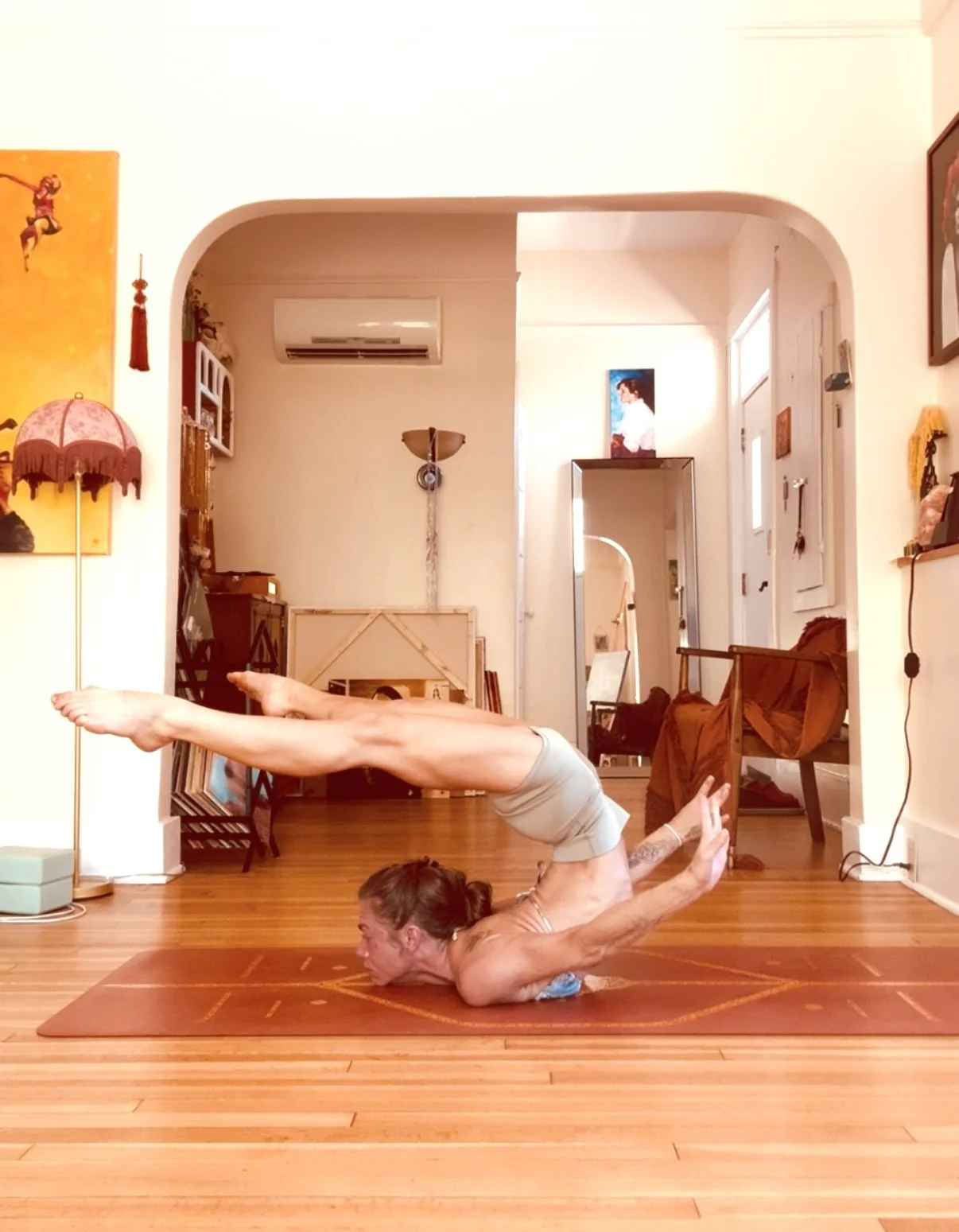 Woman practicing yoga in the dolphin pose on a yoga mat inside a living room with wooden floors, artwork on the walls, and furniture including a chair, table, and mirror.