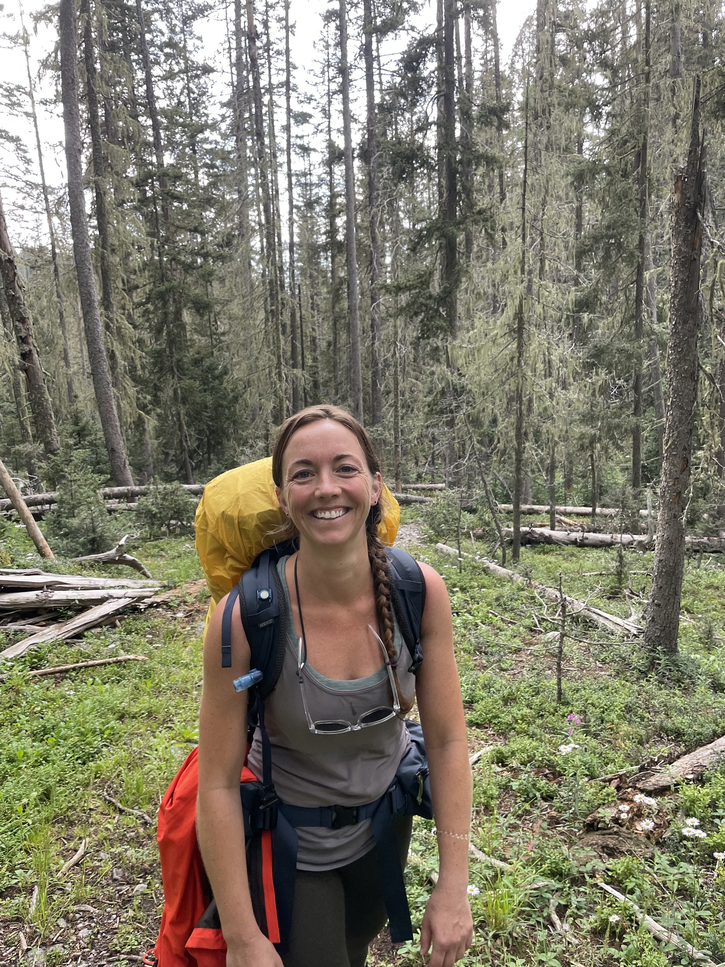 A woman with a braided hairstyle smiling while hiking in a forest with coniferous trees and small flowers on the ground, wearing a gray tank top, glasses hanging on her neck, and carrying a yellow backpack.