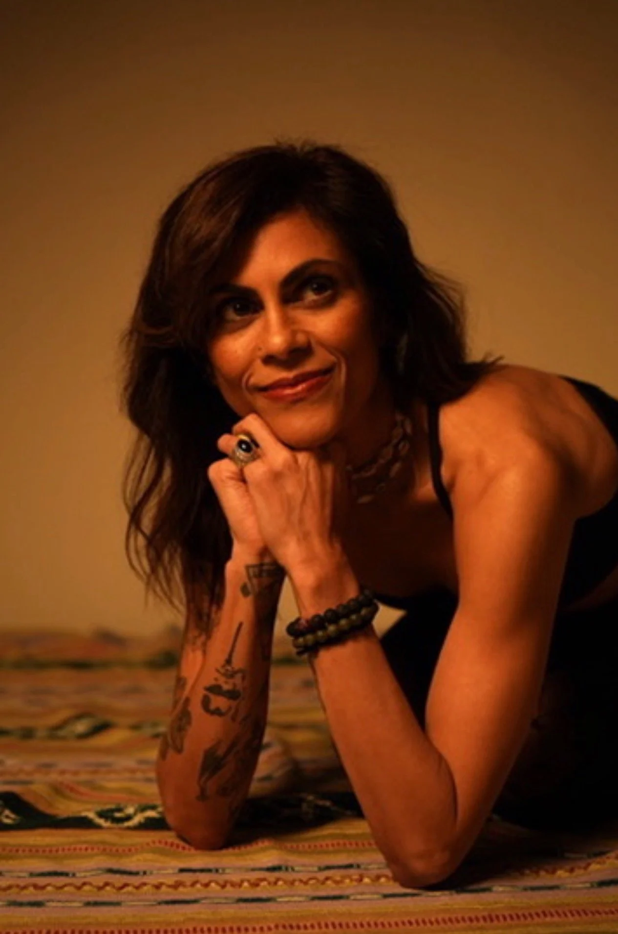 Woman with brown hair, wearing black tank top, posing on her elbows on a colorful striped rug, with her chin resting on her right hand, looking to her left.
