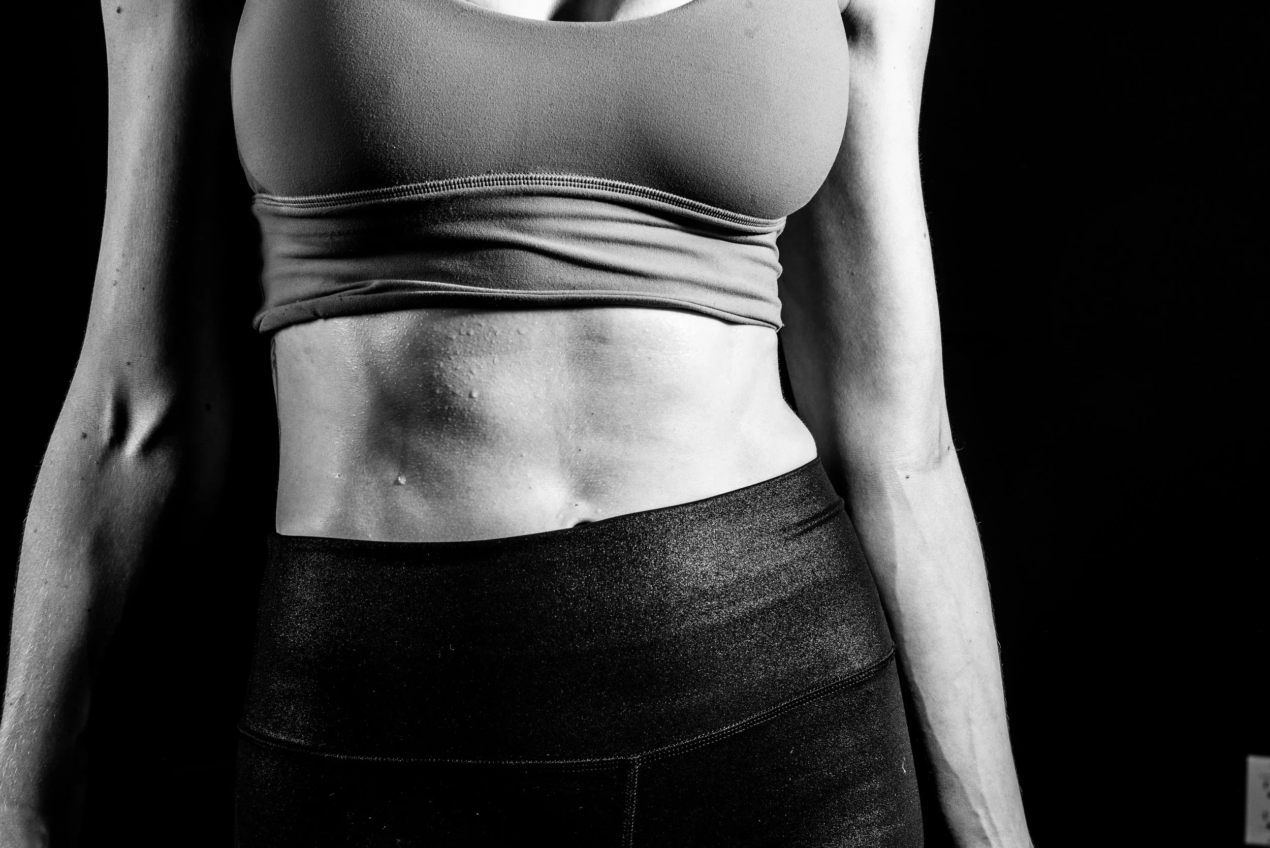Close-up of a woman’s midsection, wearing a sports bra and workout leggings, with sweat on her skin, against a black background.