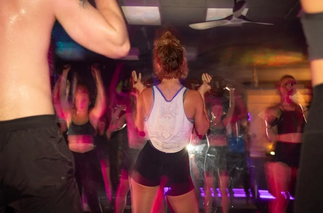 A woman with curly hair facing away, wearing a white tank top and black shorts, dancing in a club with colorful lights and other people around her.