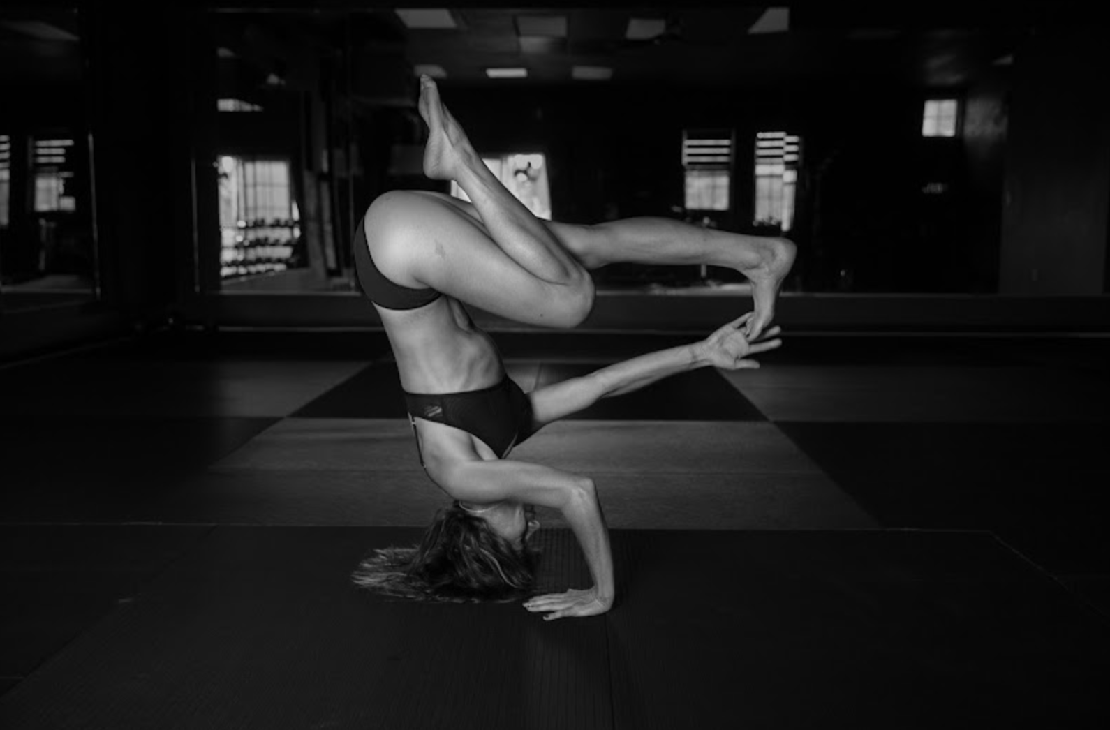 A woman practicing yoga indoors, balancing on her hands with her legs bent and lifted, captured in black and white.