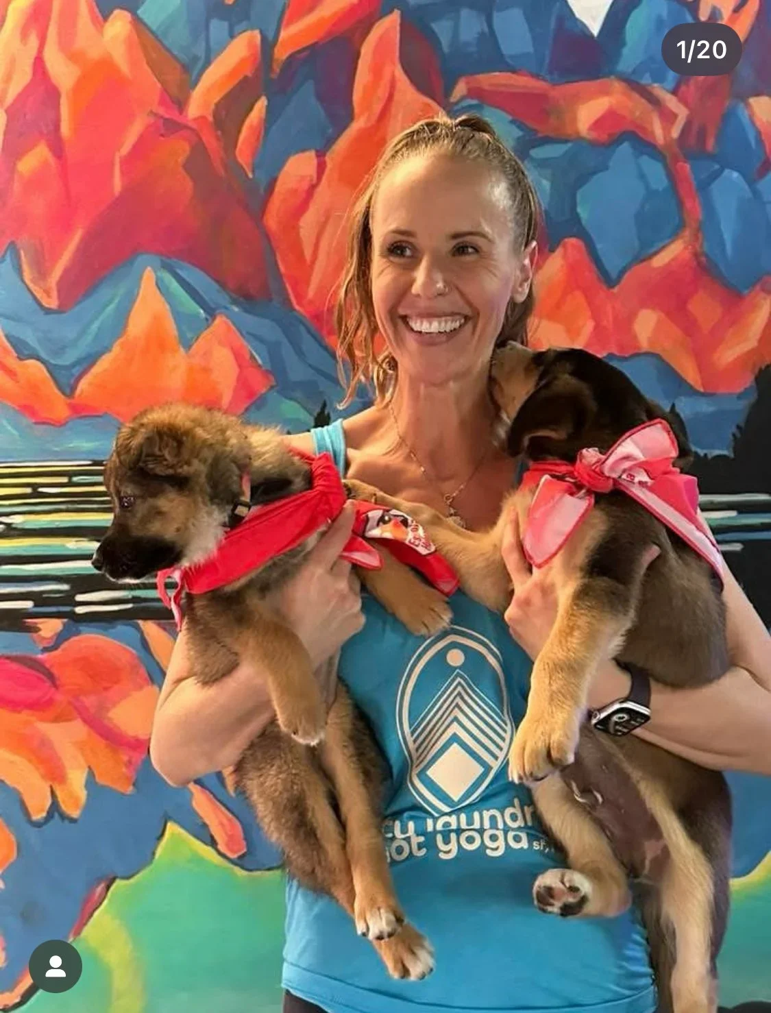 A smiling woman holding two puppies wearing red bandanas, standing in front of a colorful abstract mural.