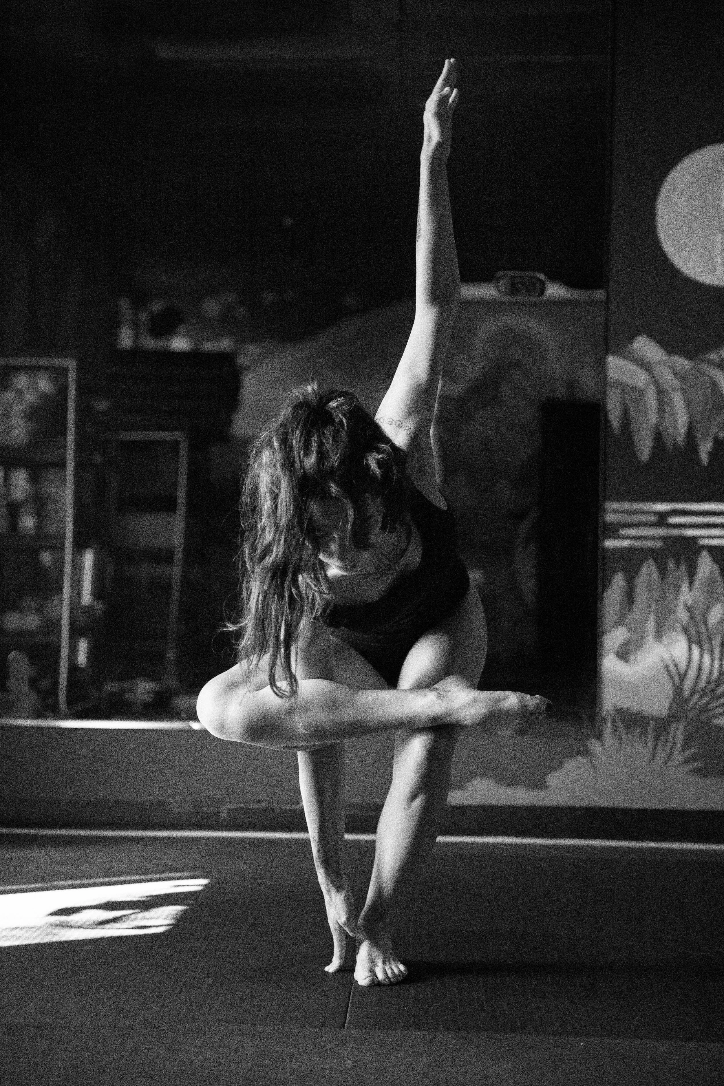 A woman practicing yoga or dance indoors, balancing on one leg with her other leg bent, and her arm extended upward. She has long, wavy hair and is wearing a sleeveless top. The background features various artworks on the walls.