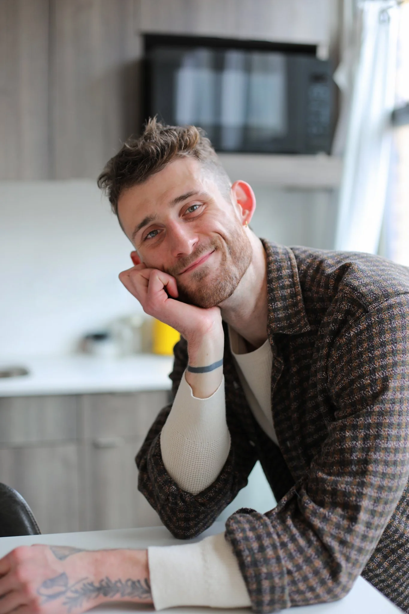 A young man with short curly hair, light skin, and a beard, resting his head on his hand, smiling at the camera. He wears a checkered shirt and has a tattoo on his forearm. The background shows a kitchen with a microwave and window.