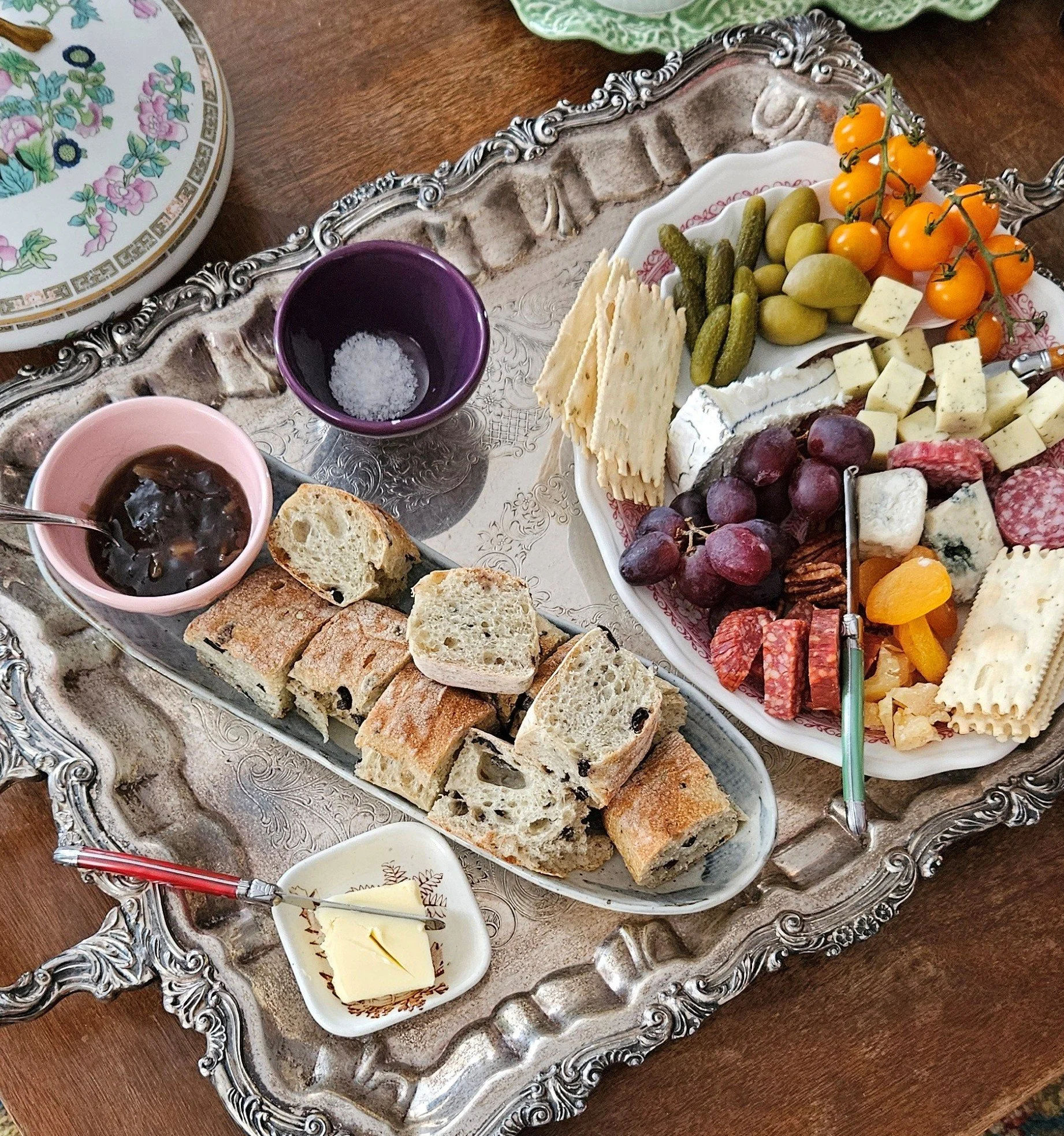 A silver ornate tray holding sliced bread, a small dish of butter, a plate of cheeses, grapes, figs, cherry tomatoes, pickles, and crackers. There are also two colorful cups, one purple and one pink, with beverages, and a small dish of jam or jelly. The table surface is wooden.