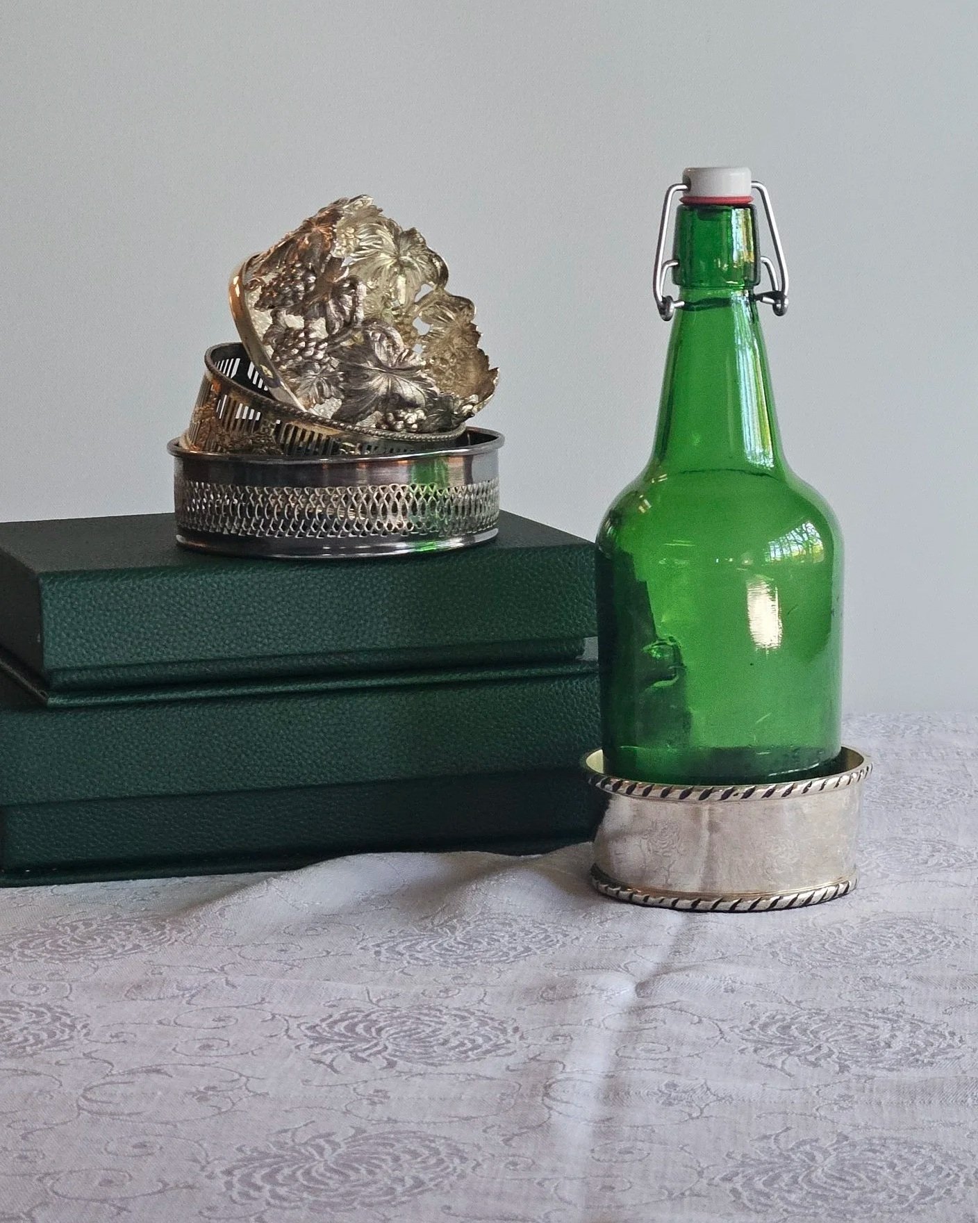 A green glass bottle with a swing top closure on a silver tray, placed on a table with a white patterned tablecloth, a green textured box, and a small metal container with intricate floral designs.