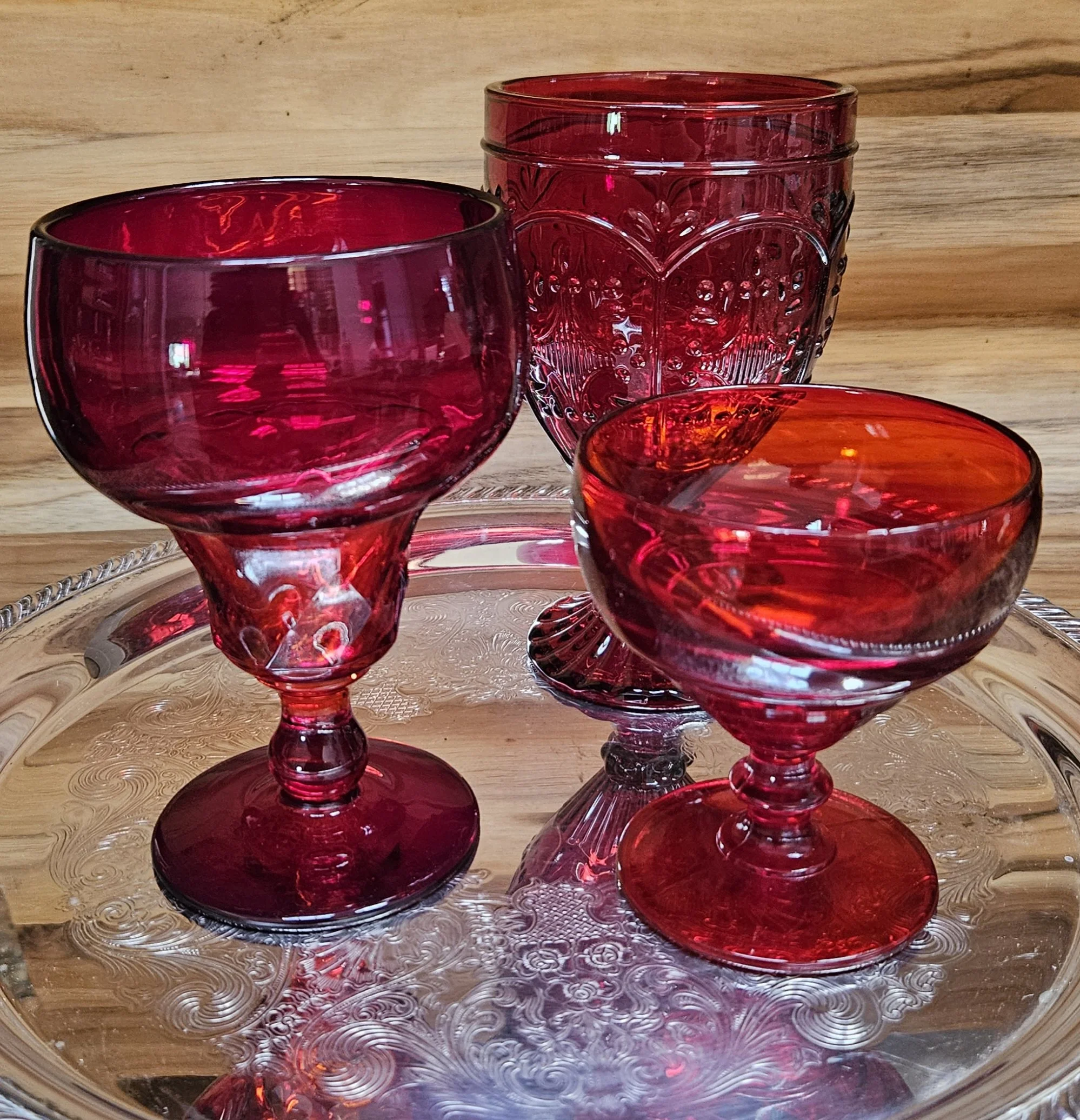 Close-up of three red glass candle holders with intricate patterns, arranged on a clear, decorative glass tray with an etched swirl design, set against a wooden background.