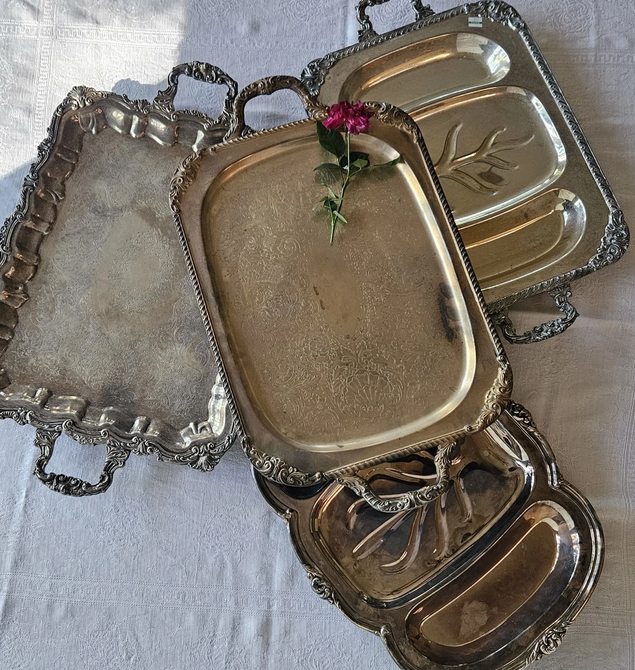 Elegant silver trays with intricate designs and a pink flower placed on one of the trays, set on a white tablecloth.