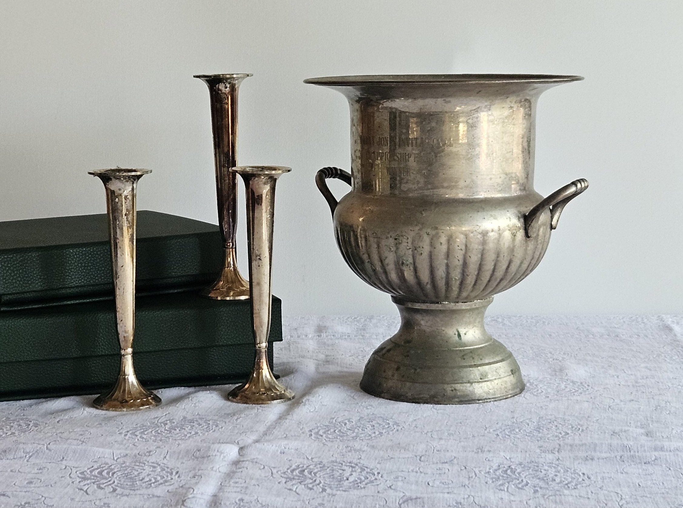 Four antique candlesticks and a silver vase arranged on a white textured surface against a plain wall, with books stacked behind.
