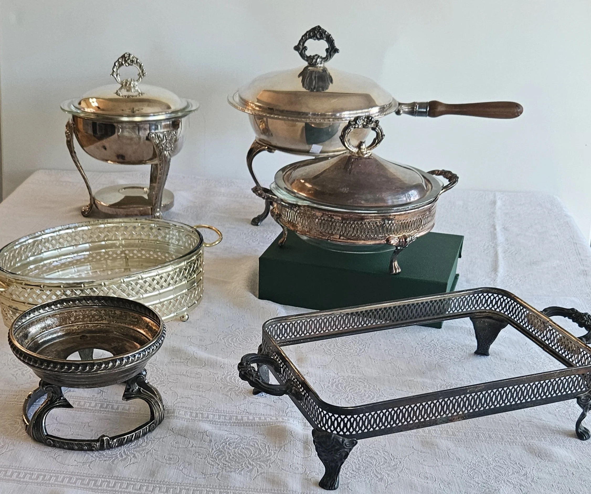 A collection of vintage silver and metal serving bowls, trays, and stands displayed on a white tablecloth, with some pieces featuring ornate handles and decorative legs.