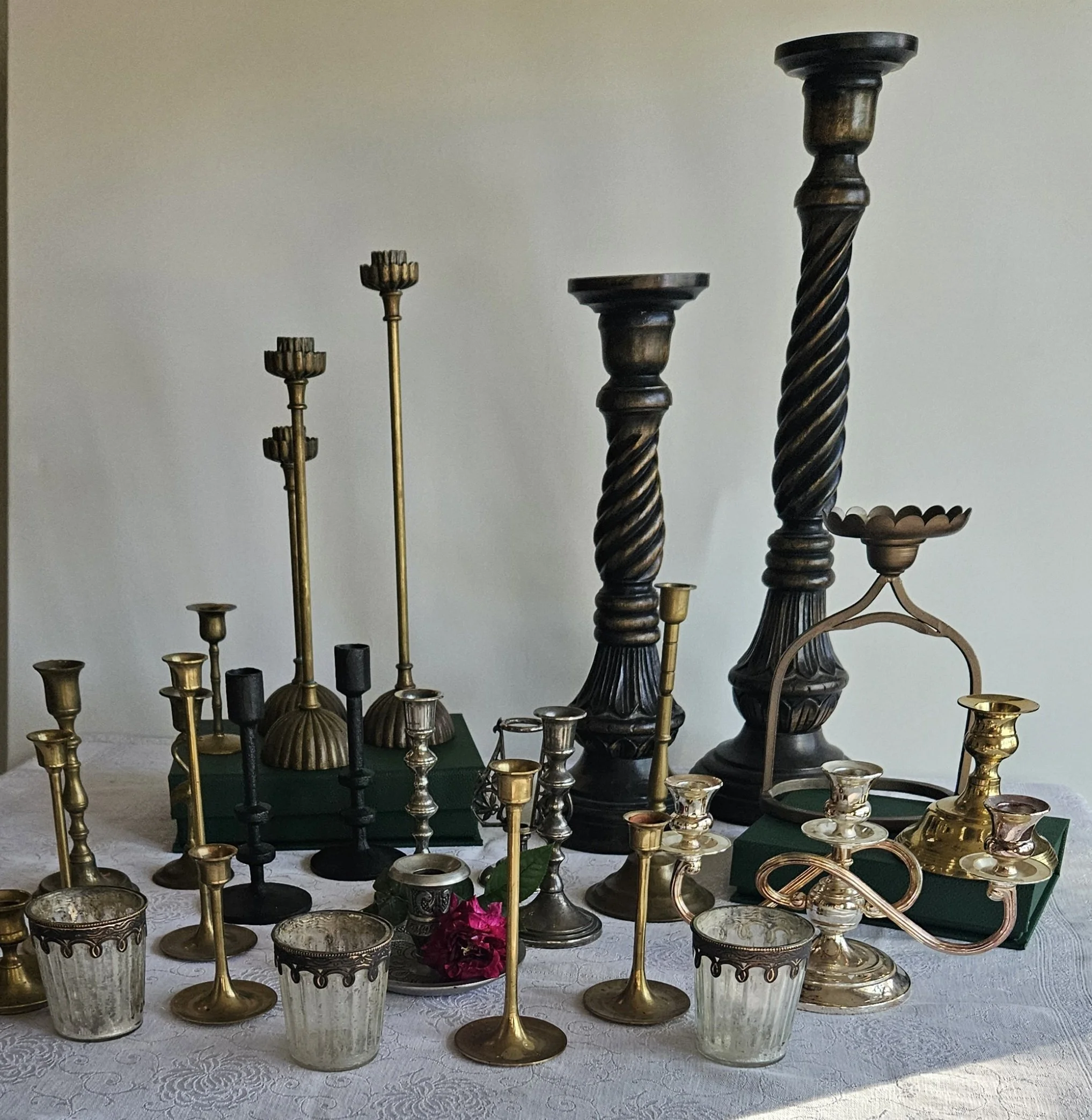Various decorative candle holders made of metal and glass arranged on a table with a white lace tablecloth. The candle holders are of different heights and designs, with some being taller and others shorter, and feature intricate patterns and twisted designs.