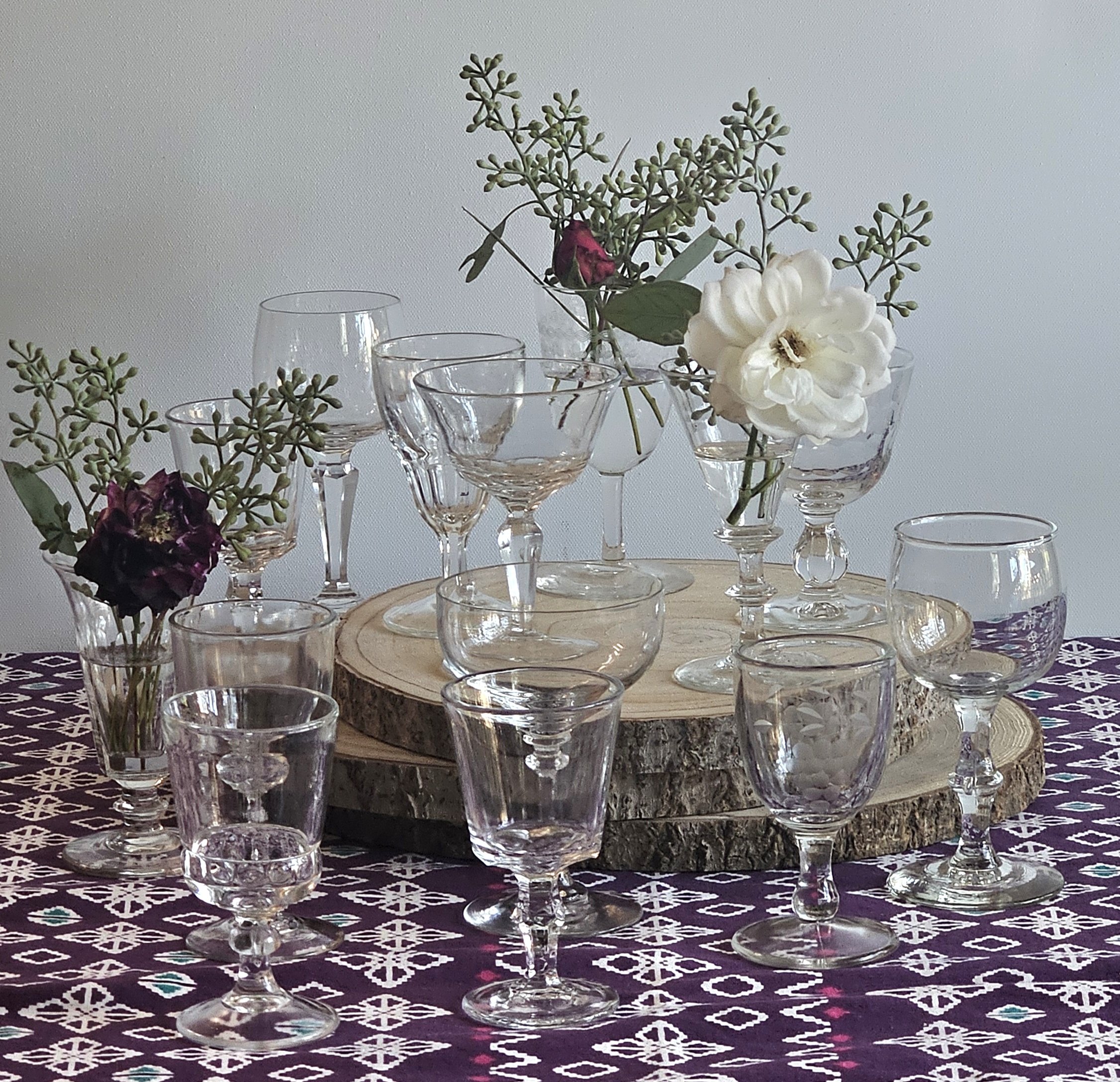 A floral arrangement in a glass vase decorated with clear glass cups, featuring cream-colored flowers and green foliage, placed on a wooden tray on a patterned tablecloth.