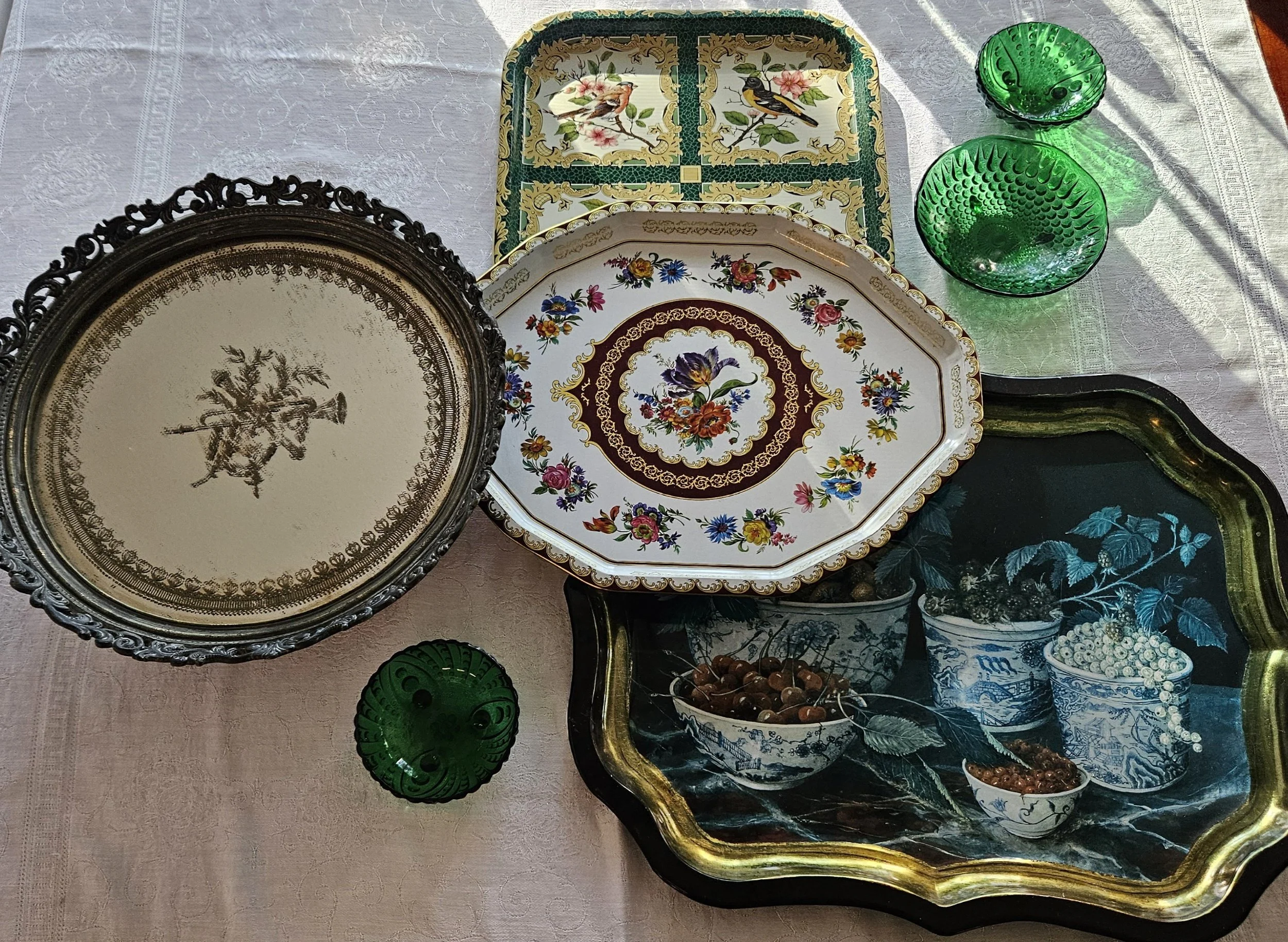 Various decorative dishes and glasses on a table, including floral, bird, and landscape designs, with two green glass bowls and a silver-framed mirror.