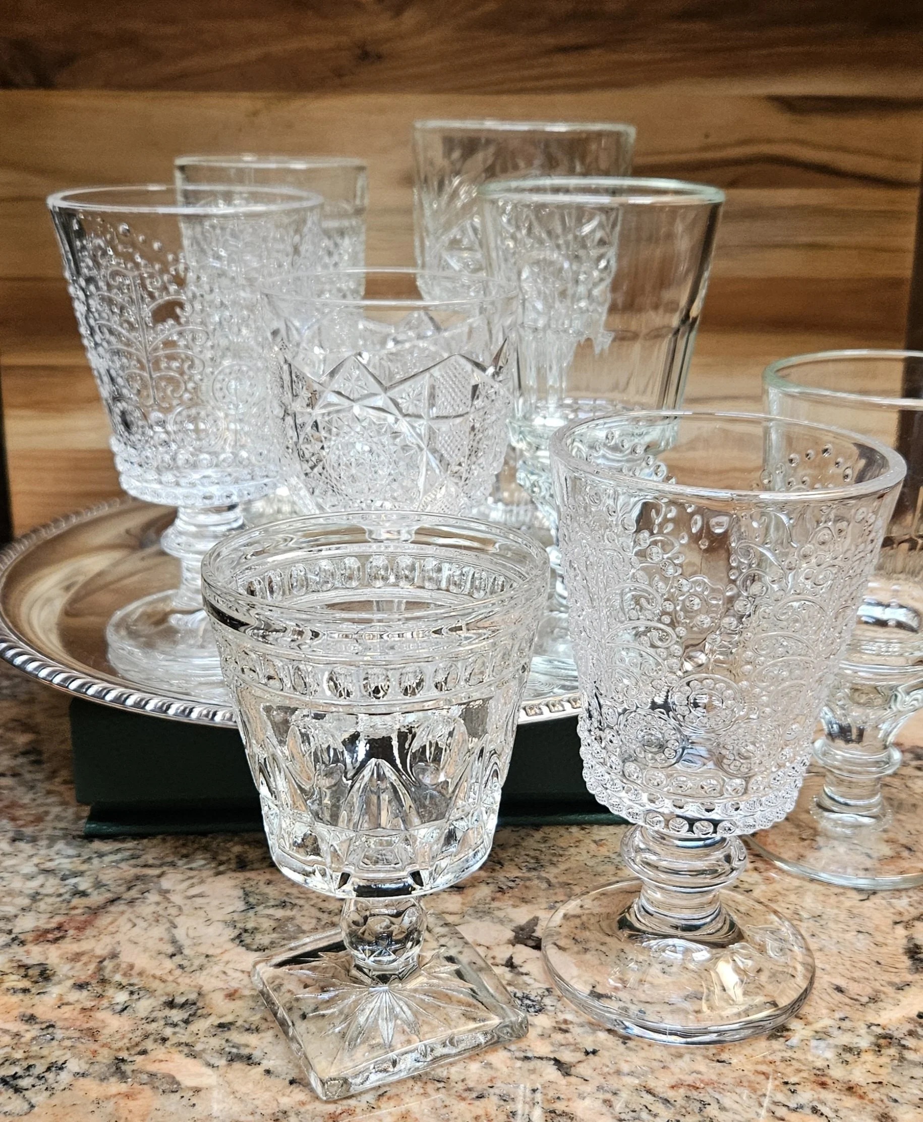 A collection of clear, intricately cut glassware includes various glasses with decorative patterns, placed on a silver tray on a granite countertop with a wooden background.