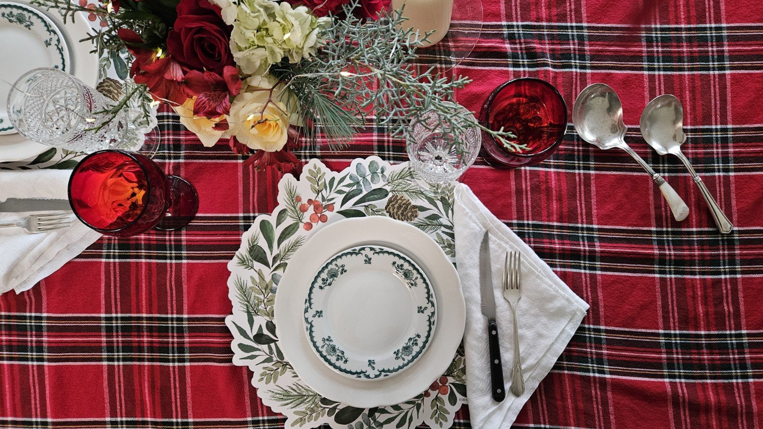 A holiday table setting with a red plaid tablecloth, white and green decorative plates, silverware, red and clear glasses, a floral centerpiece with greenery and red and white flowers, and holiday-themed napkins.