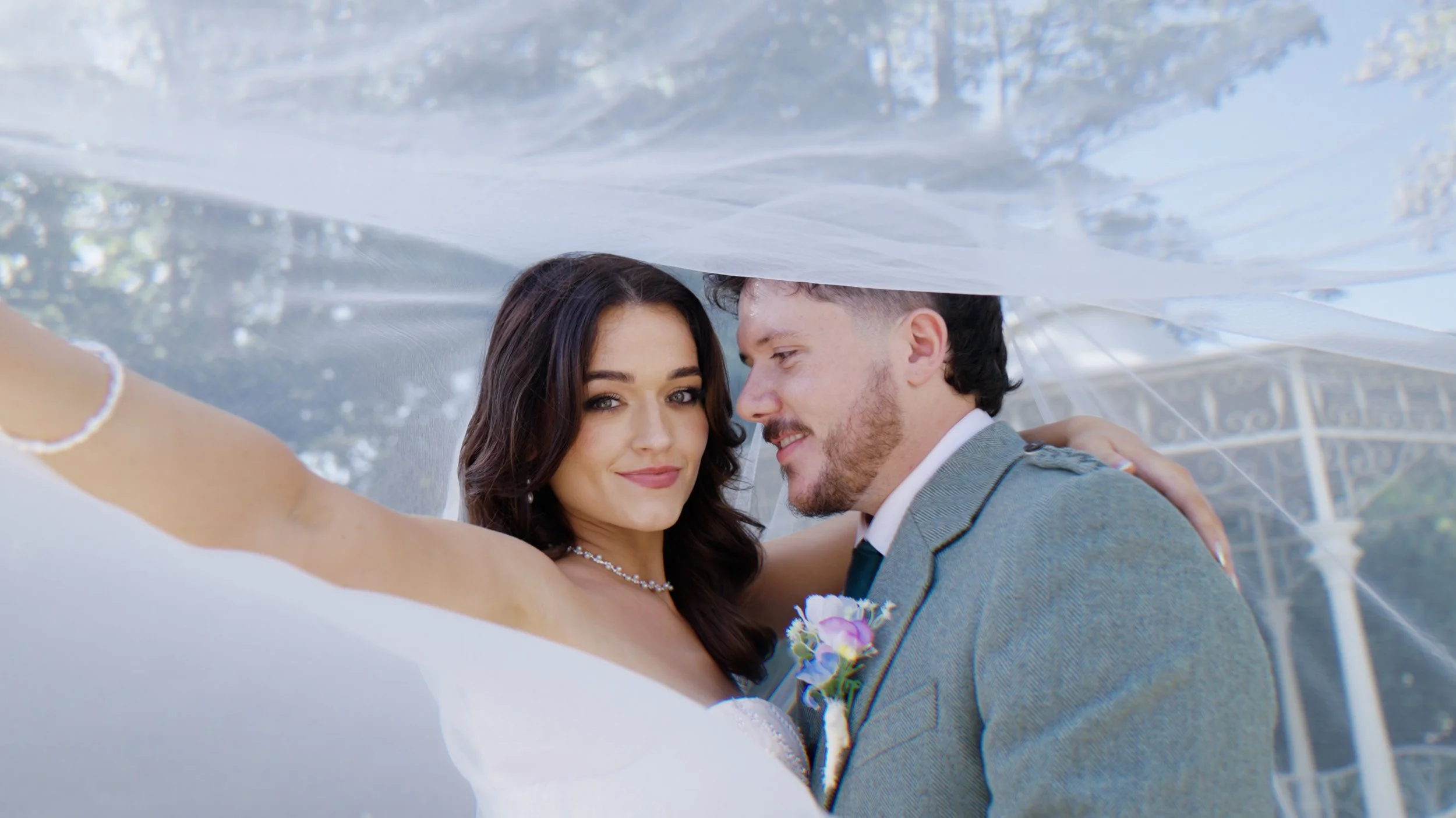 A happy bride and groom sharing a kiss under a white wedding veil outdoors, with trees in the background.
