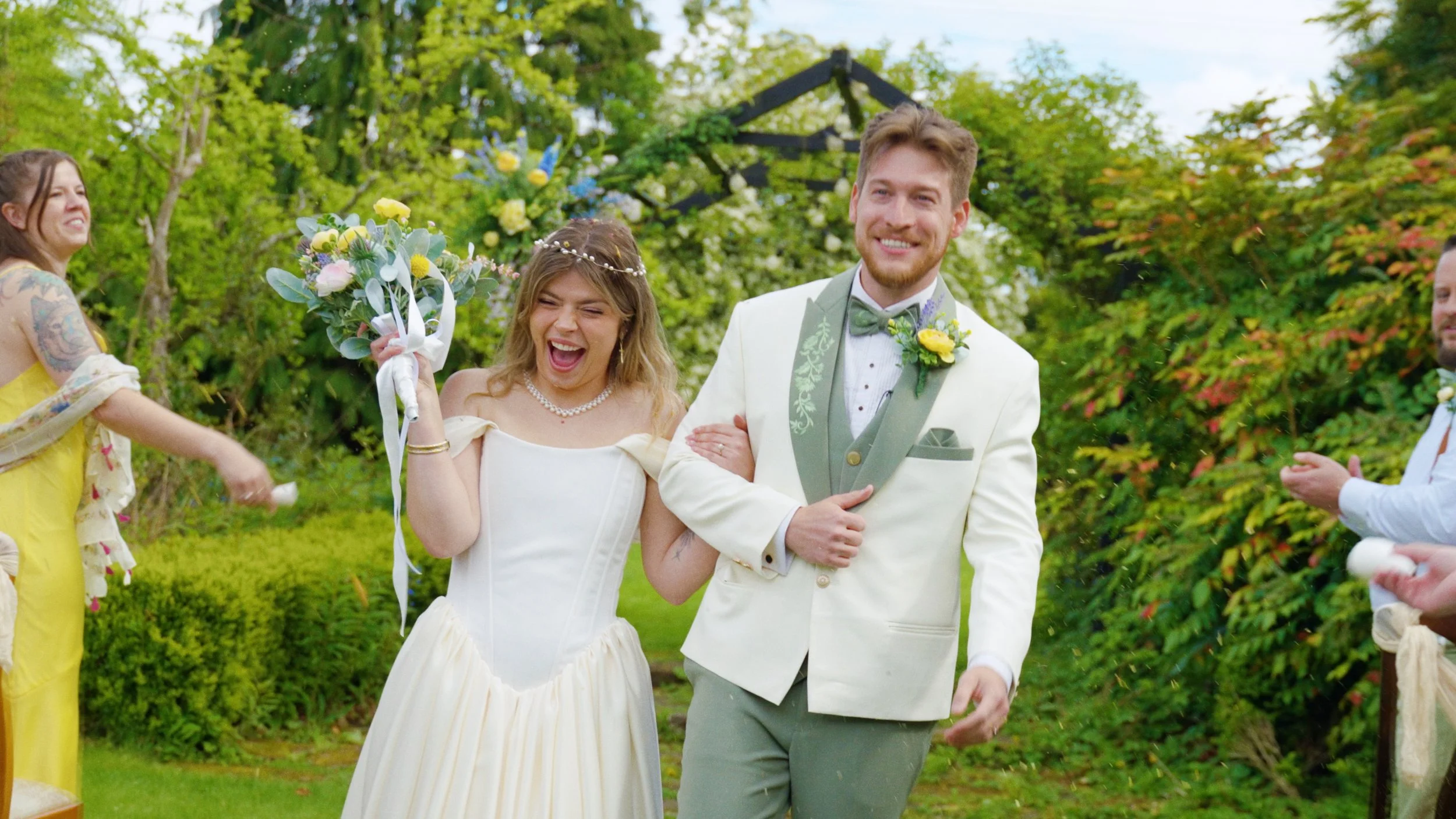 A bride and groom smiling at their wedding outdoors, surrounded by green trees and colorful flowers, with guests celebrating around them.