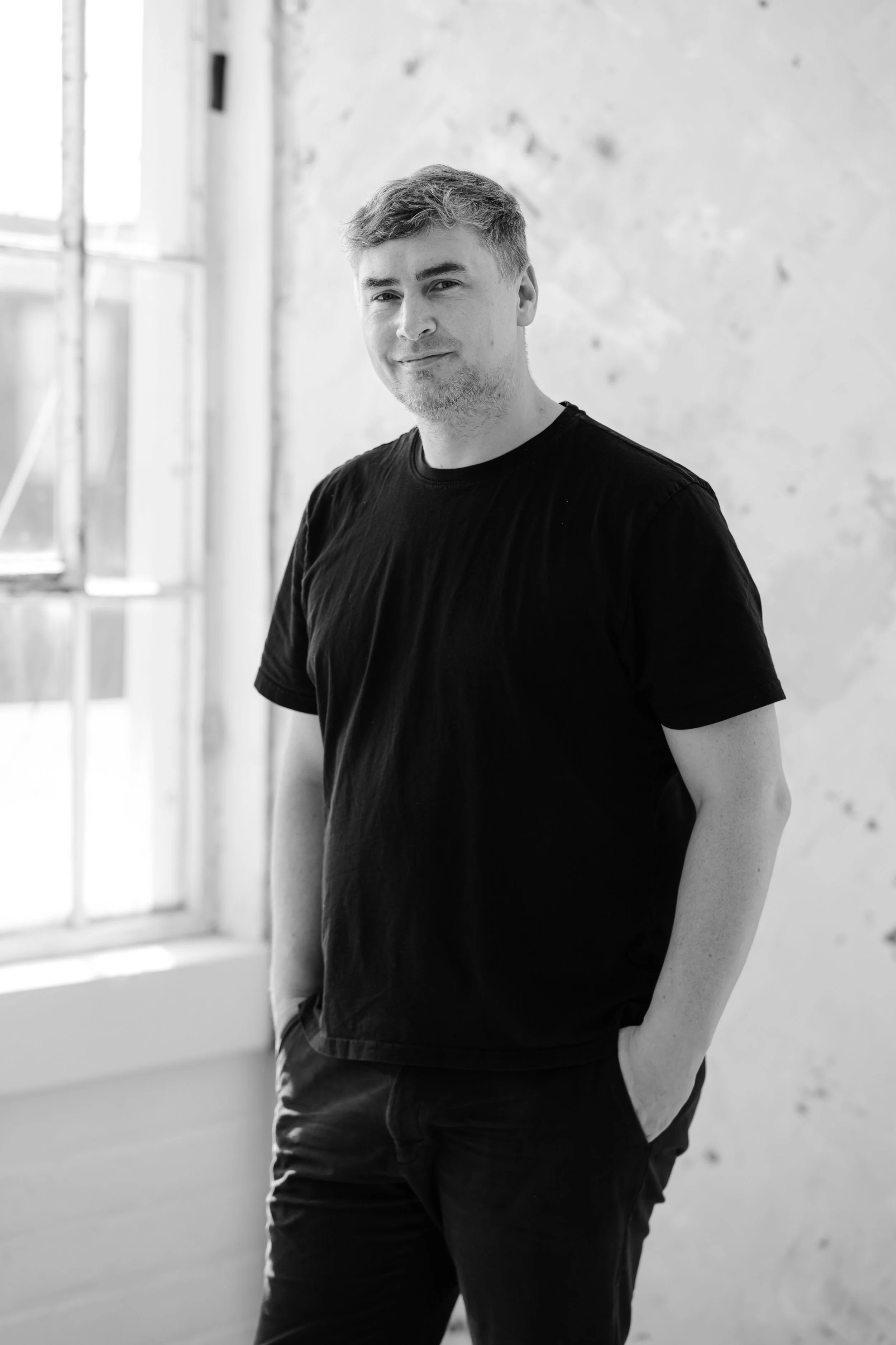 Black and white photo of a young man wearing a casual t-shirt, standing indoors next to a window with sunlight, with a textured wall in the background.