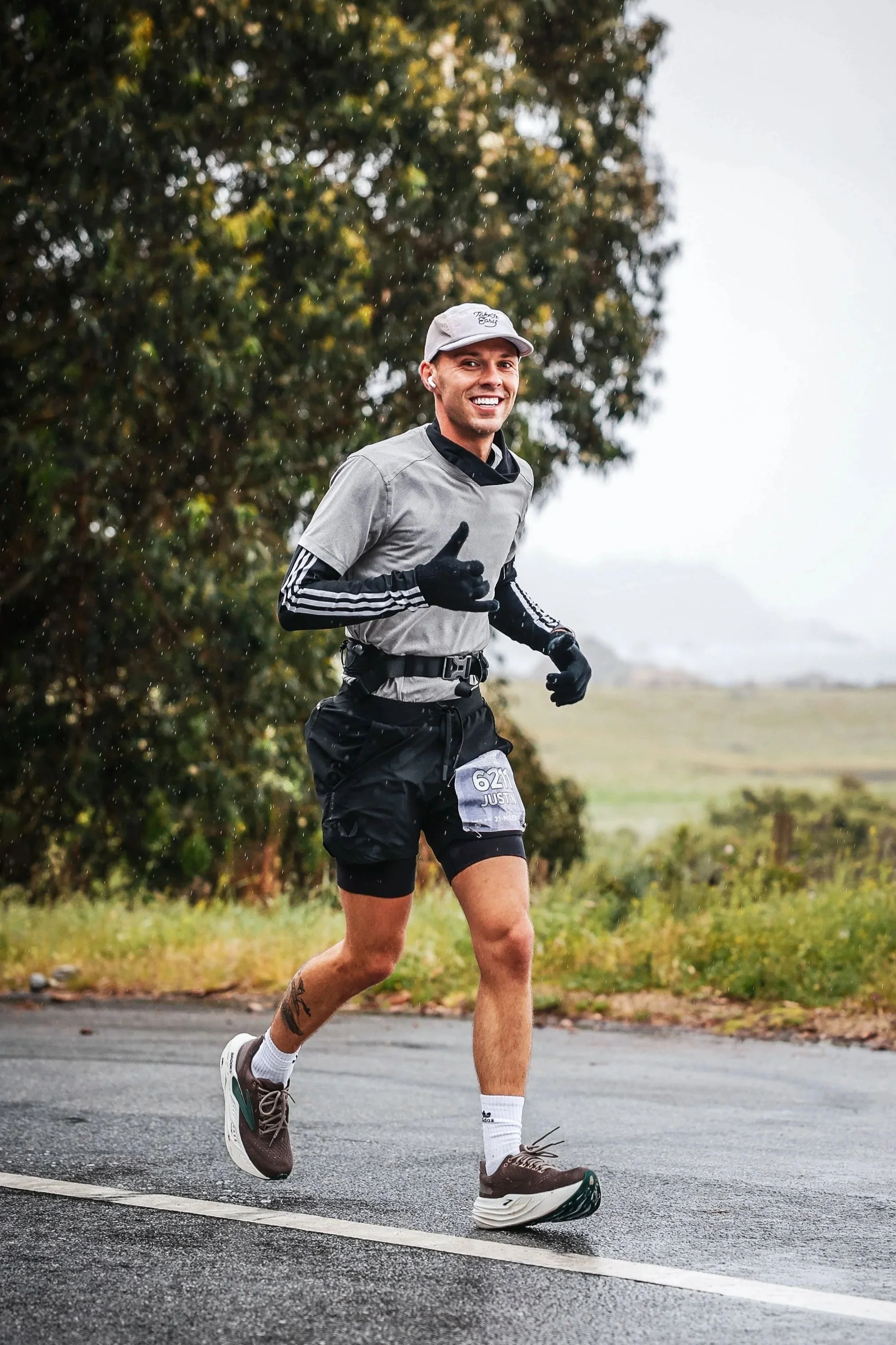 A man jogging outdoors on a paved road, wearing a gray athletic shirt, black shorts, a white cap, and sneakers, smiling and giving a thumbs-up.