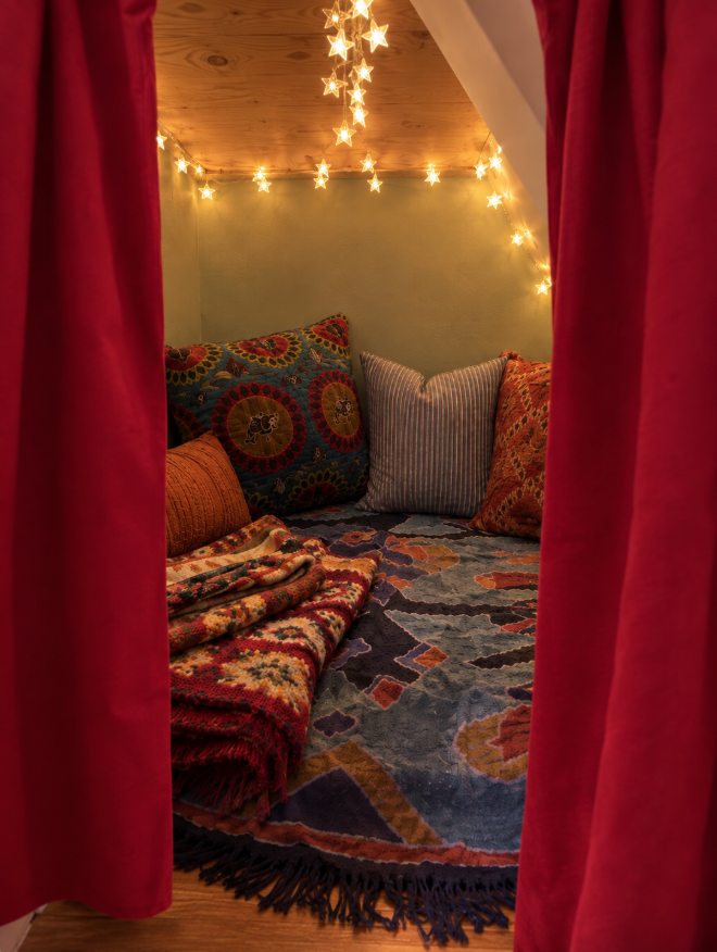 Cozy under-the-stairs nook with a red curtain, string lights and layered pillows and blankets