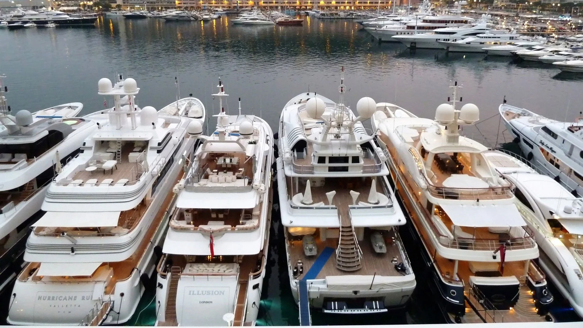 Multiple luxury yachts docked at a marina during evening with city lights reflected in the water.