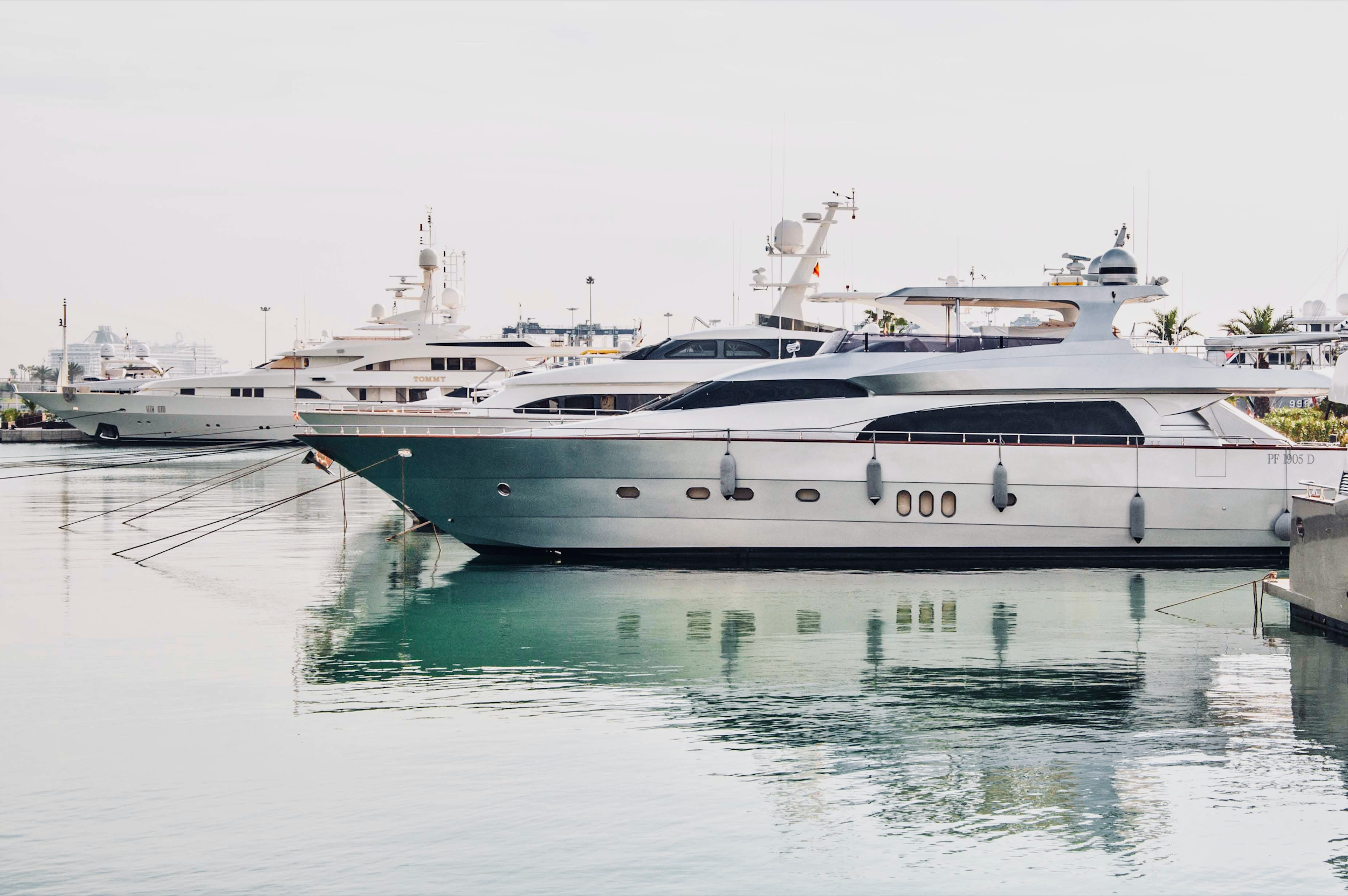 Multiple large luxury yachts docked at a marina with calm water, some palm trees, and modern buildings in the background.