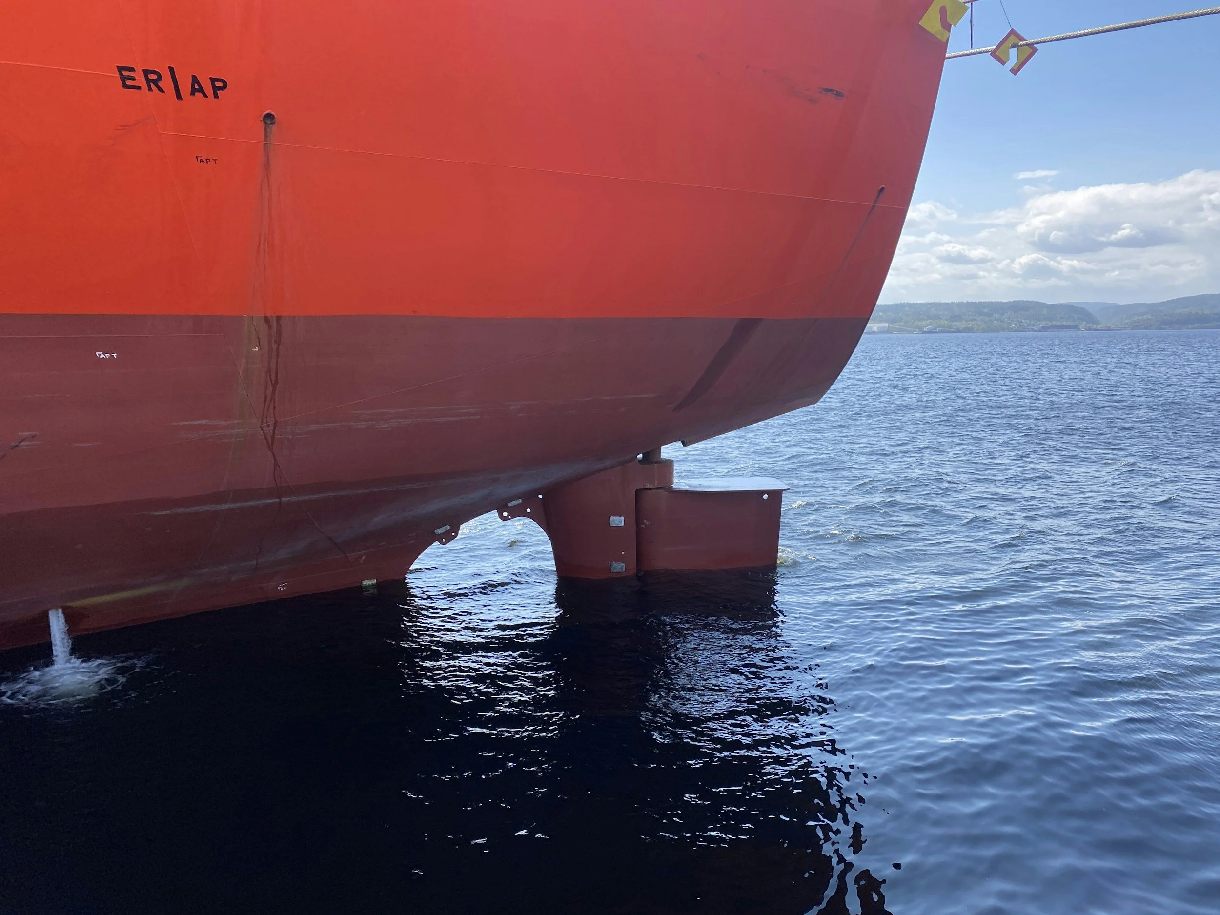 Close-up of the bow of a large red ship floating on the water under a blue sky with some clouds, with land visible in the background.