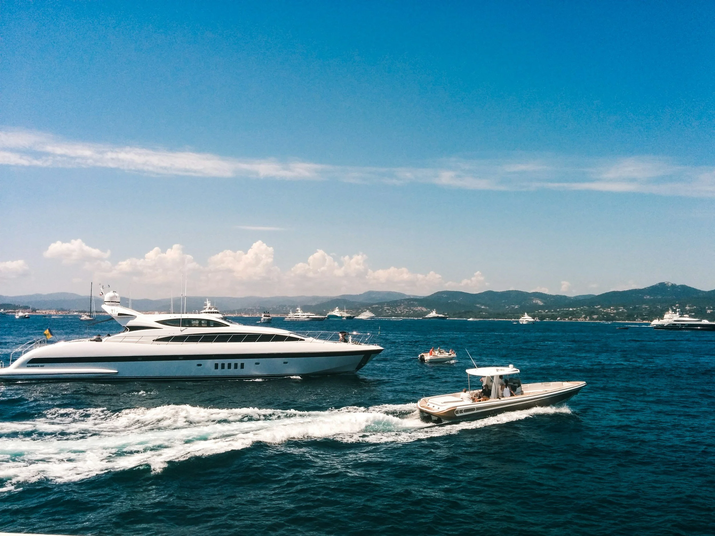 Several yachts and boats cruising on the water with mountainous landscape and partly cloudy blue sky in the background.