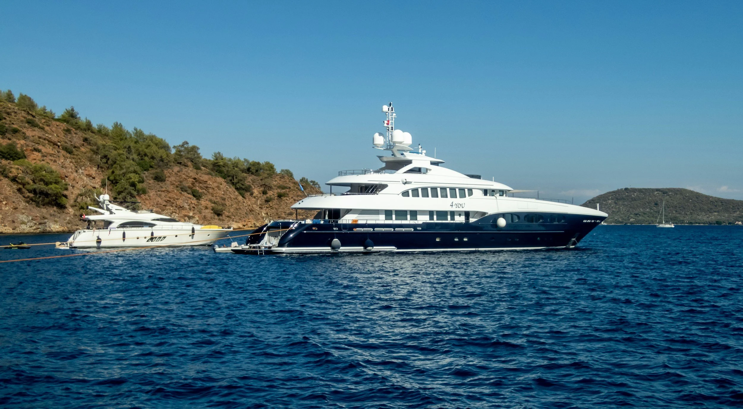 A large white yacht anchored in a body of water near a rocky hillside covered with green trees, under a clear blue sky.