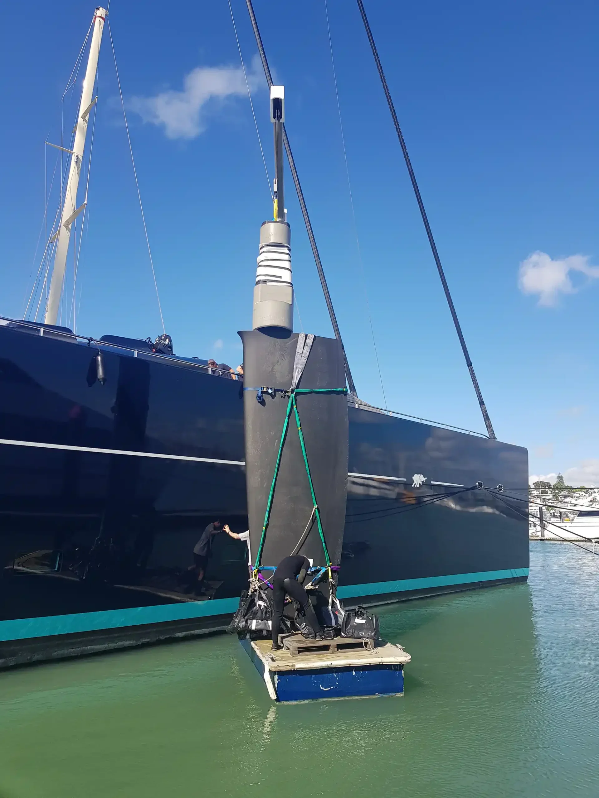 A large black sailing yacht docked at a marina, with a person standing on a small floating platform in front of it, and another person near the boat's bow. The sky is blue with a few clouds.