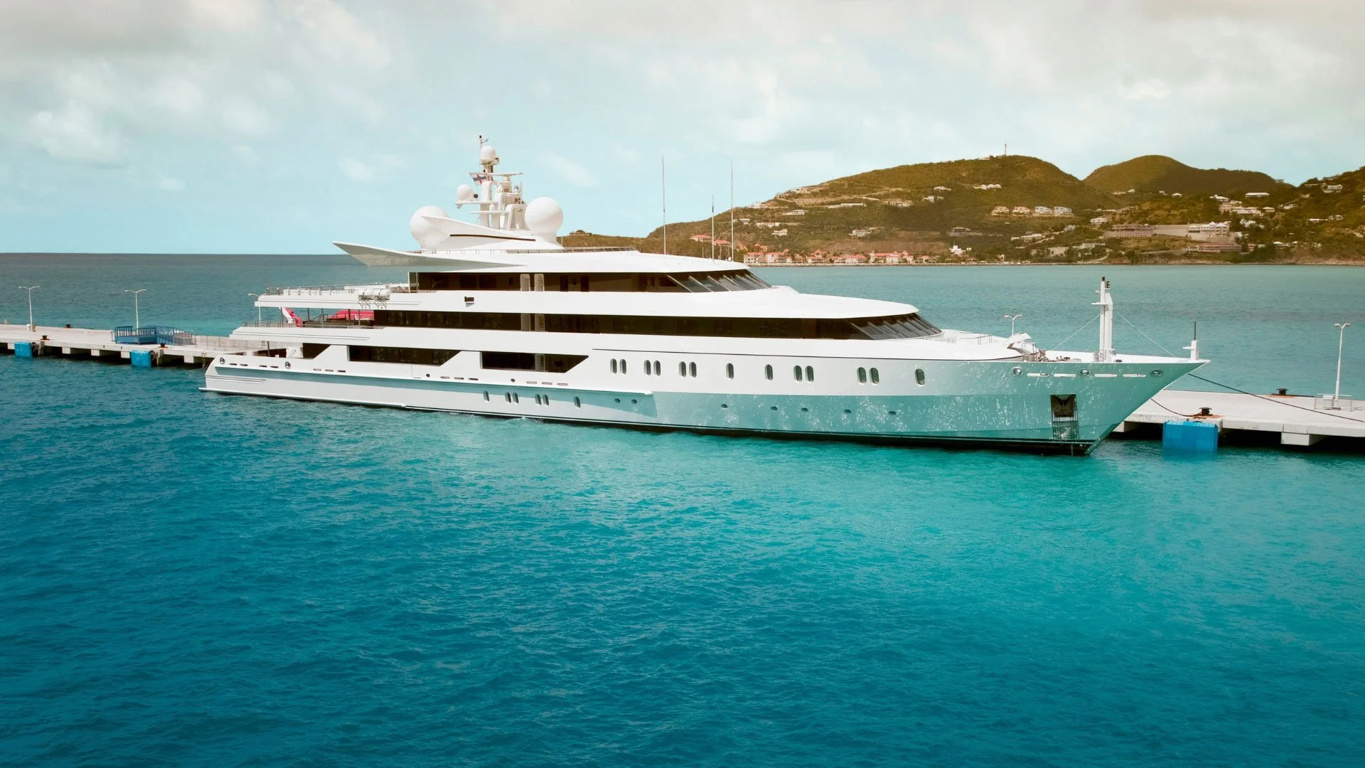 A large white yacht docked at a pier on a clear day, with a hilly island and houses in the background.