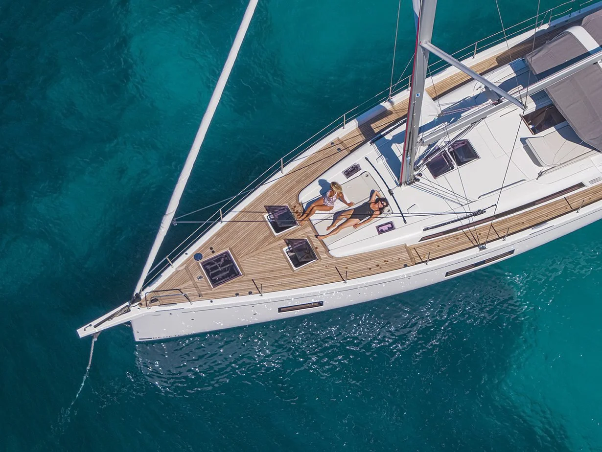 An aerial view of two women relaxing on the deck of a white sailboat in clear blue water.