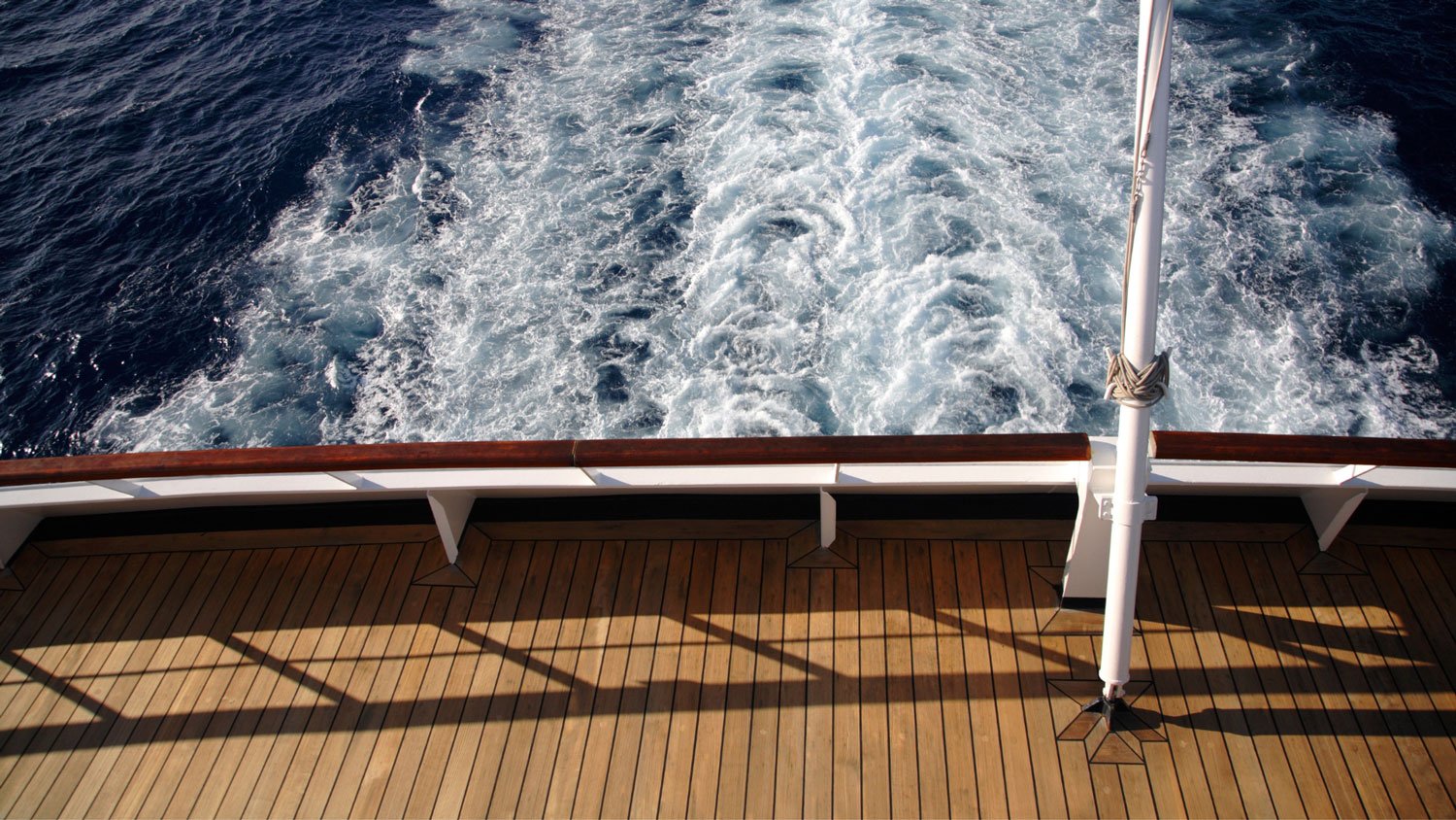 View from a boat showing a wooden deck, a metal pole with a rope, and white foamy wake in the ocean.