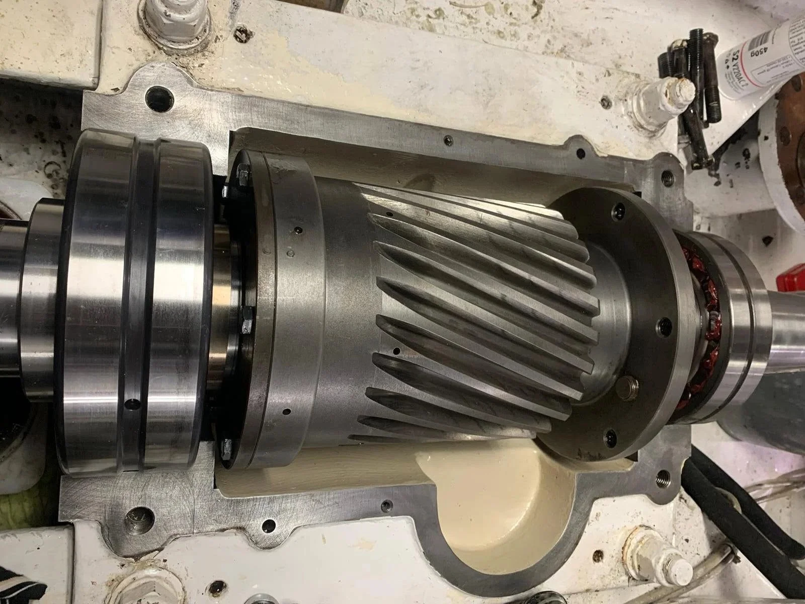Close-up of a machinery part with a large gear, a smaller pulley, and a metal casing with visible bolts, situated on a workbench with various tools and containers around.