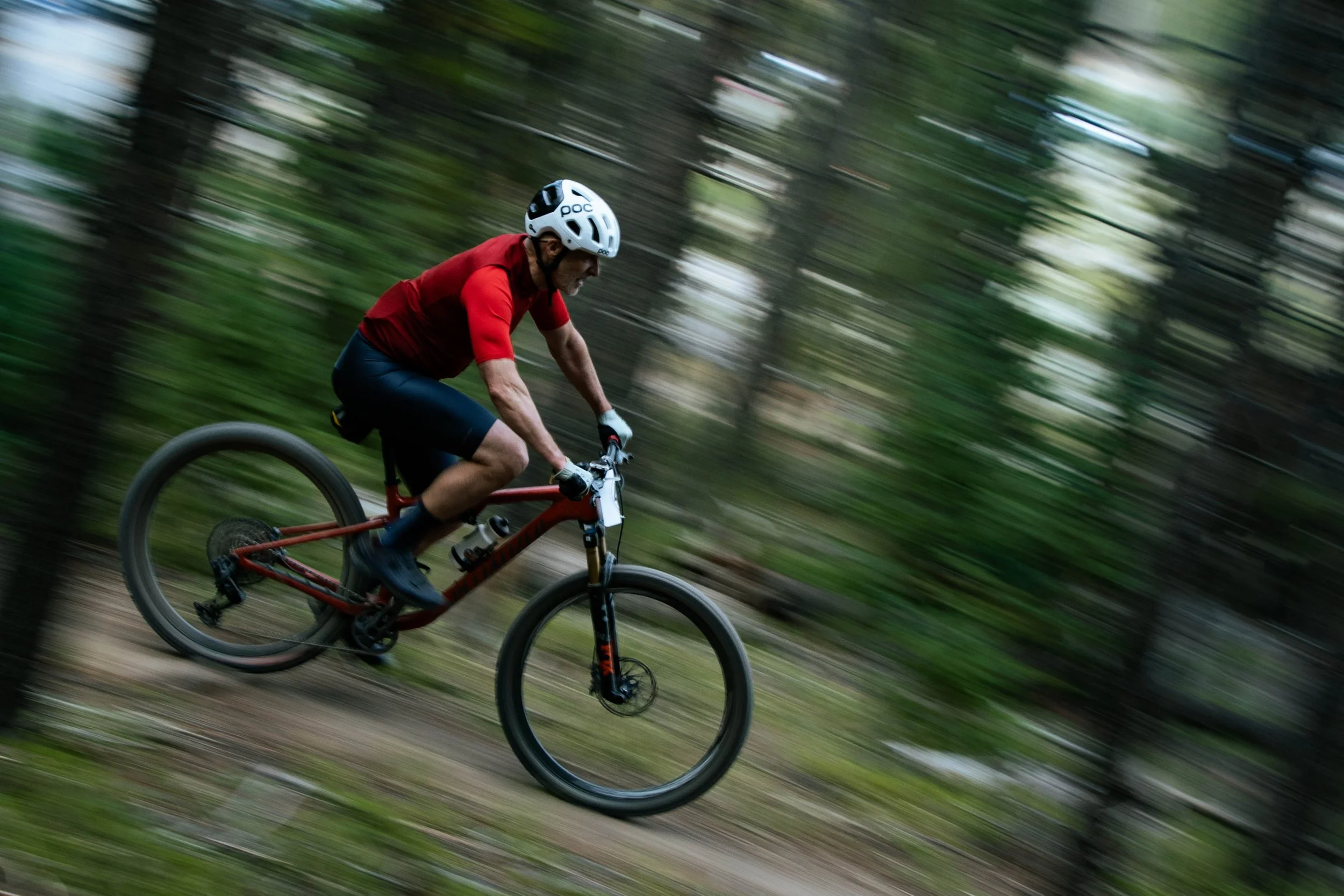 A man wearing a white helmet, red t-shirt, and riding a mountain bike through a forest trail with motion blur.
