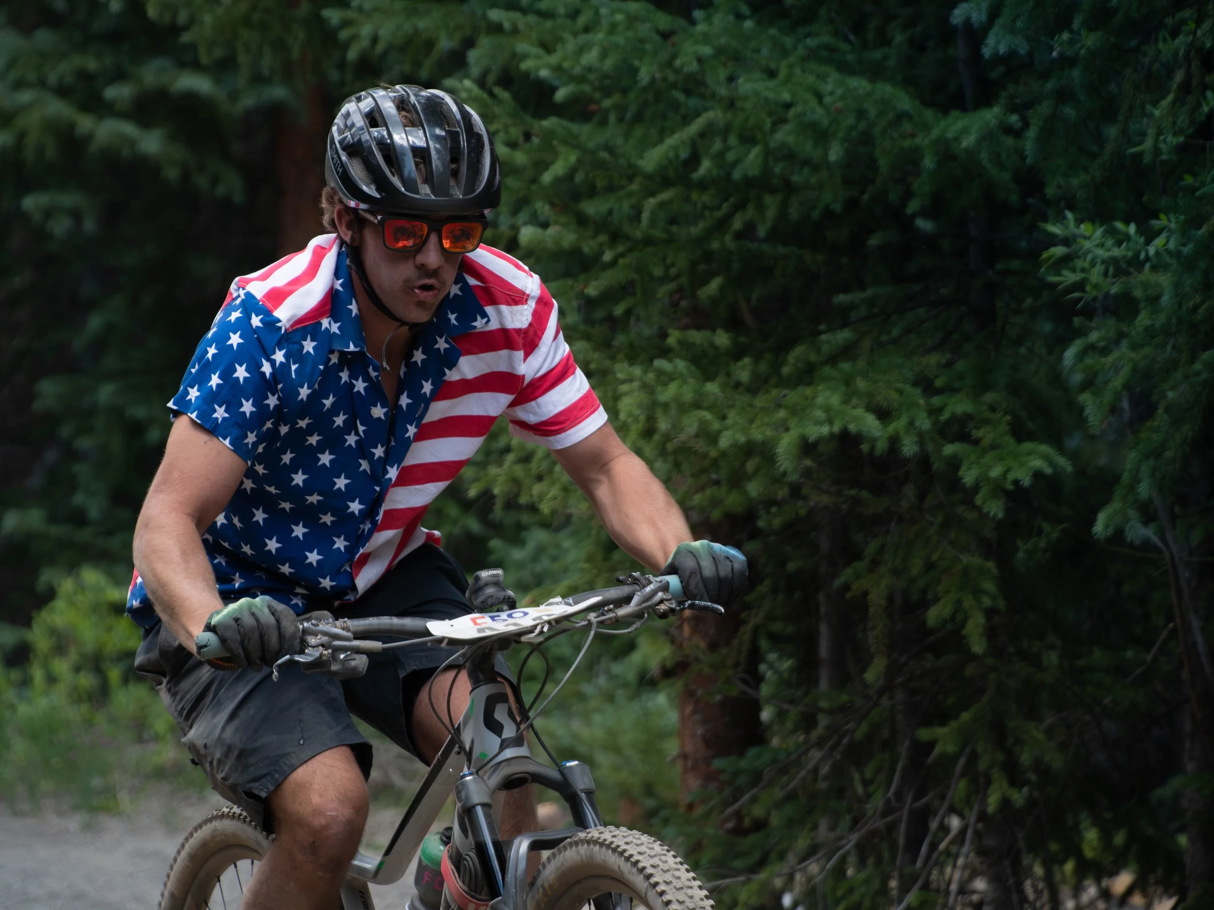A man riding a mountain bike on a trail surrounded by lush green trees, wearing a patriotic American flag shirt, a helmet, and sunglasses.