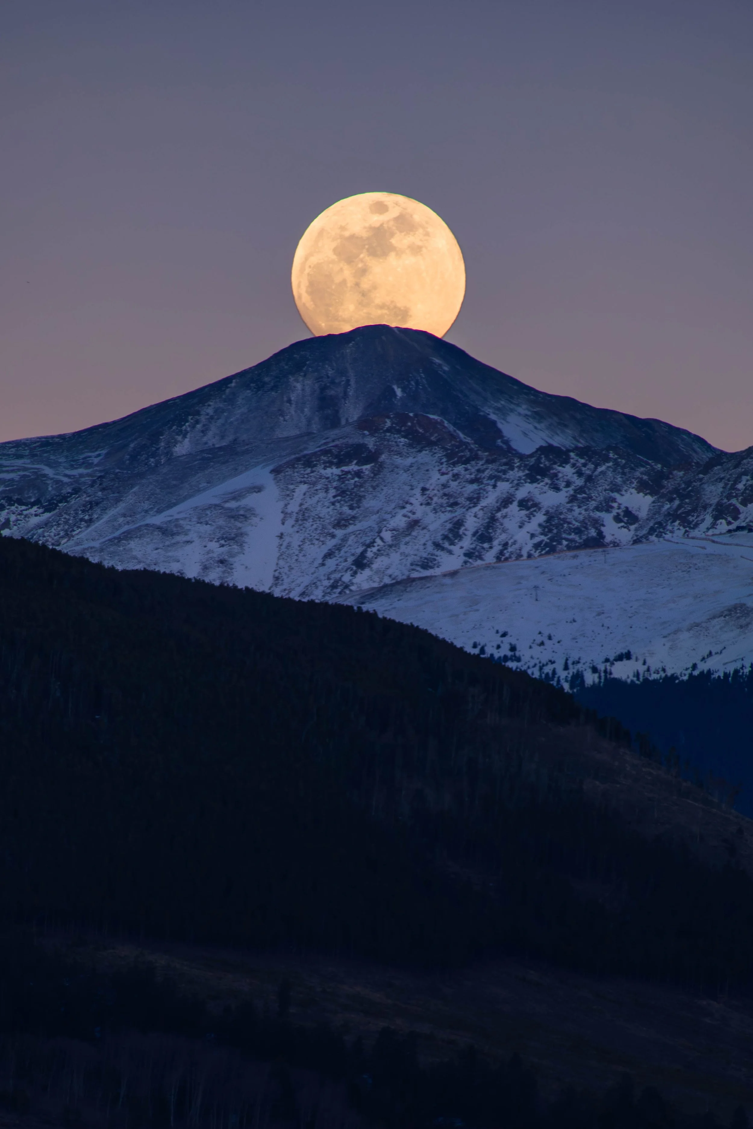 A full moon rising behind a snow-capped mountain at dusk or dawn.