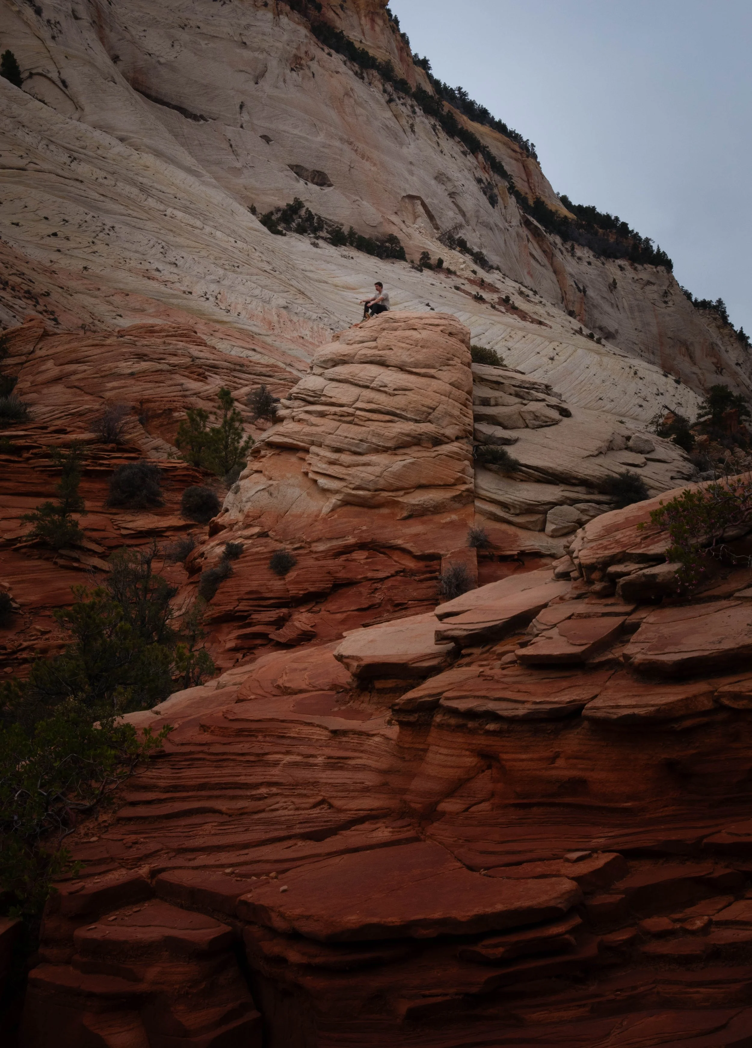 A man sitting on top of a large red rock formation in a desert landscape with layered rock formations and sparse vegetation, under an overcast sky.