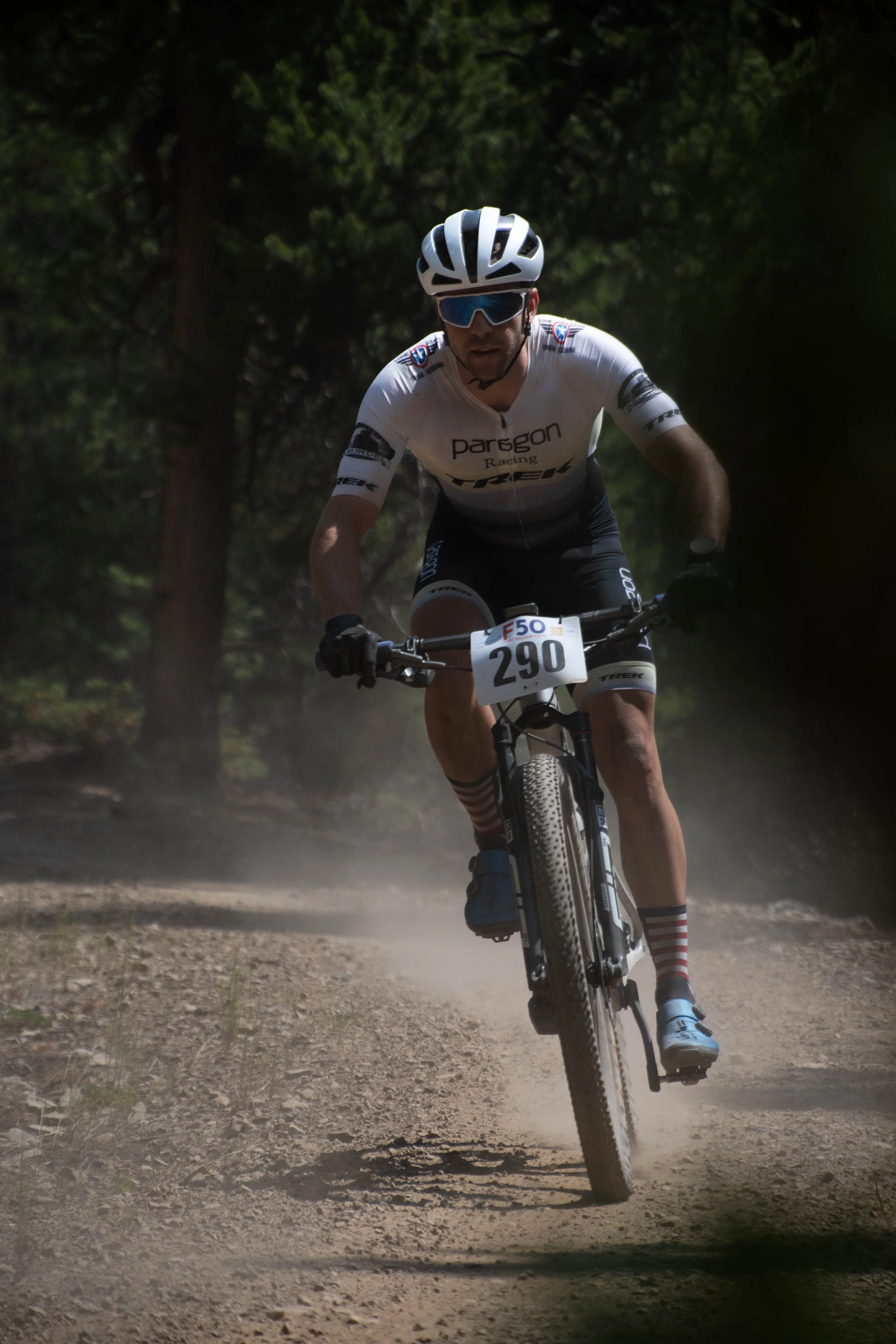 A cyclist wearing a white helmet, sunglasses, and a black and white cycling outfit riding a mountain bike on a dirt trail in a forest.