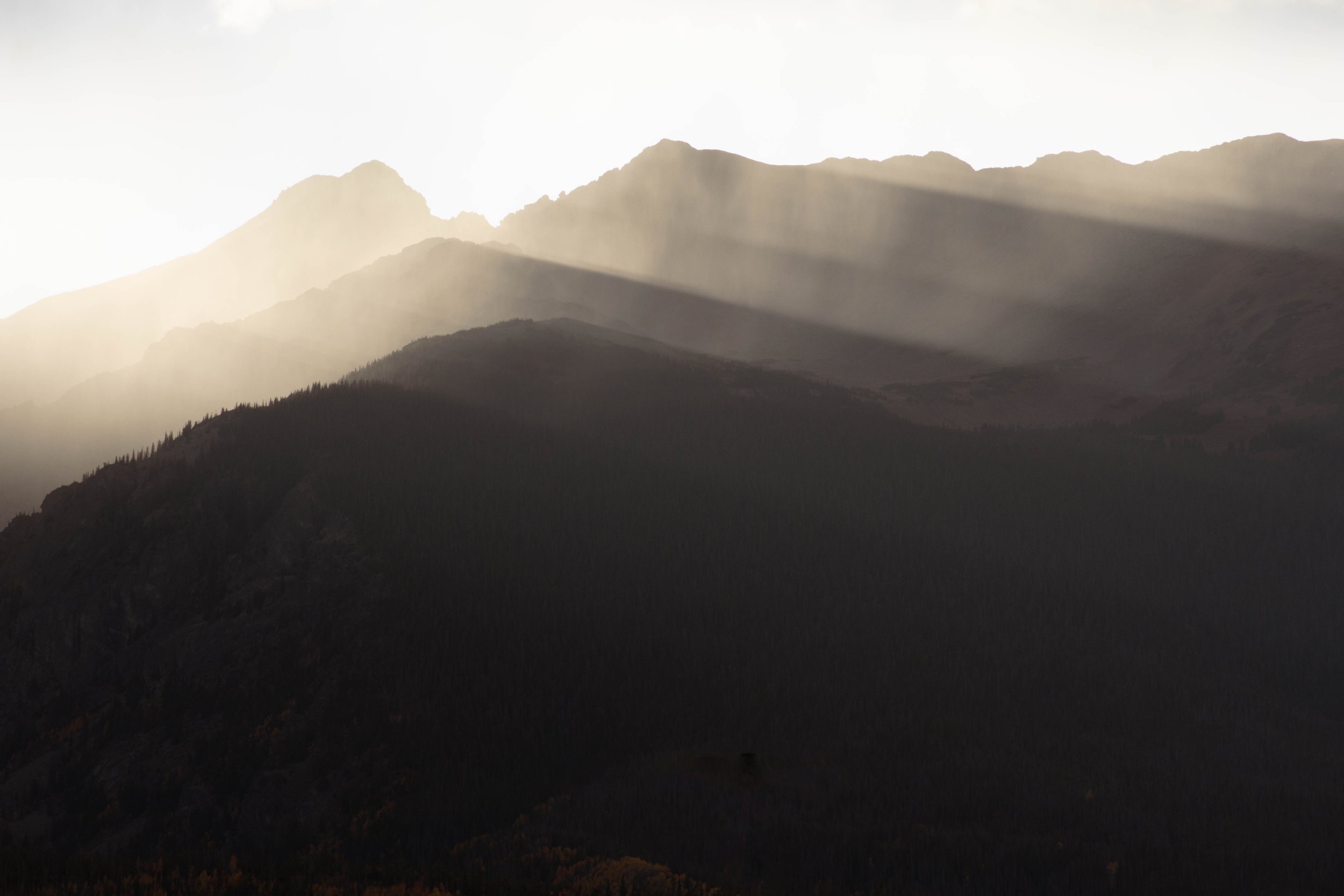 Sun rays shining through the mountains with a silhouette of forested hills in the foreground.