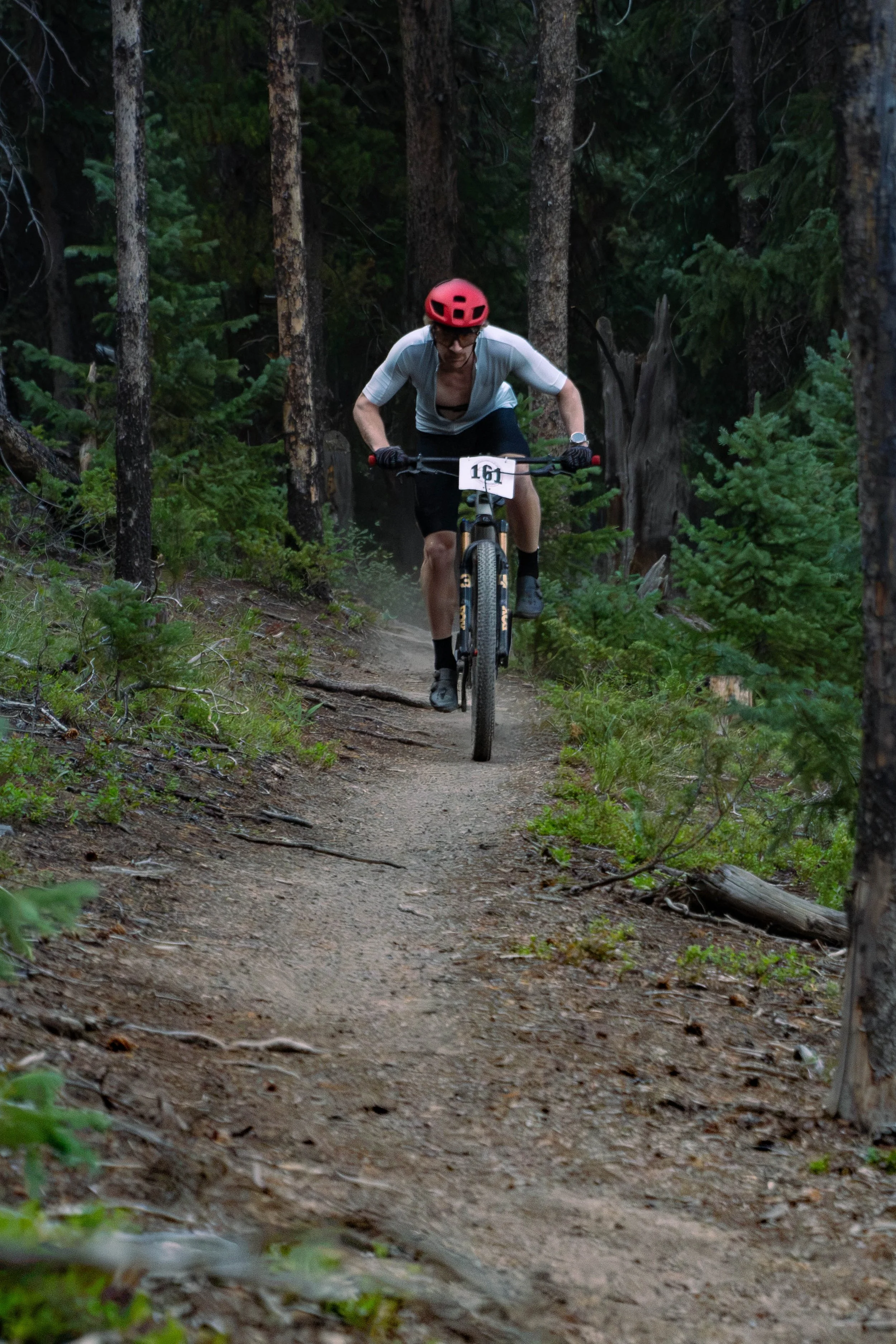 A cyclist wearing a red helmet and athletic clothing riding a mountain bike on a narrow dirt trail through a dense forest with tall trees and green foliage.