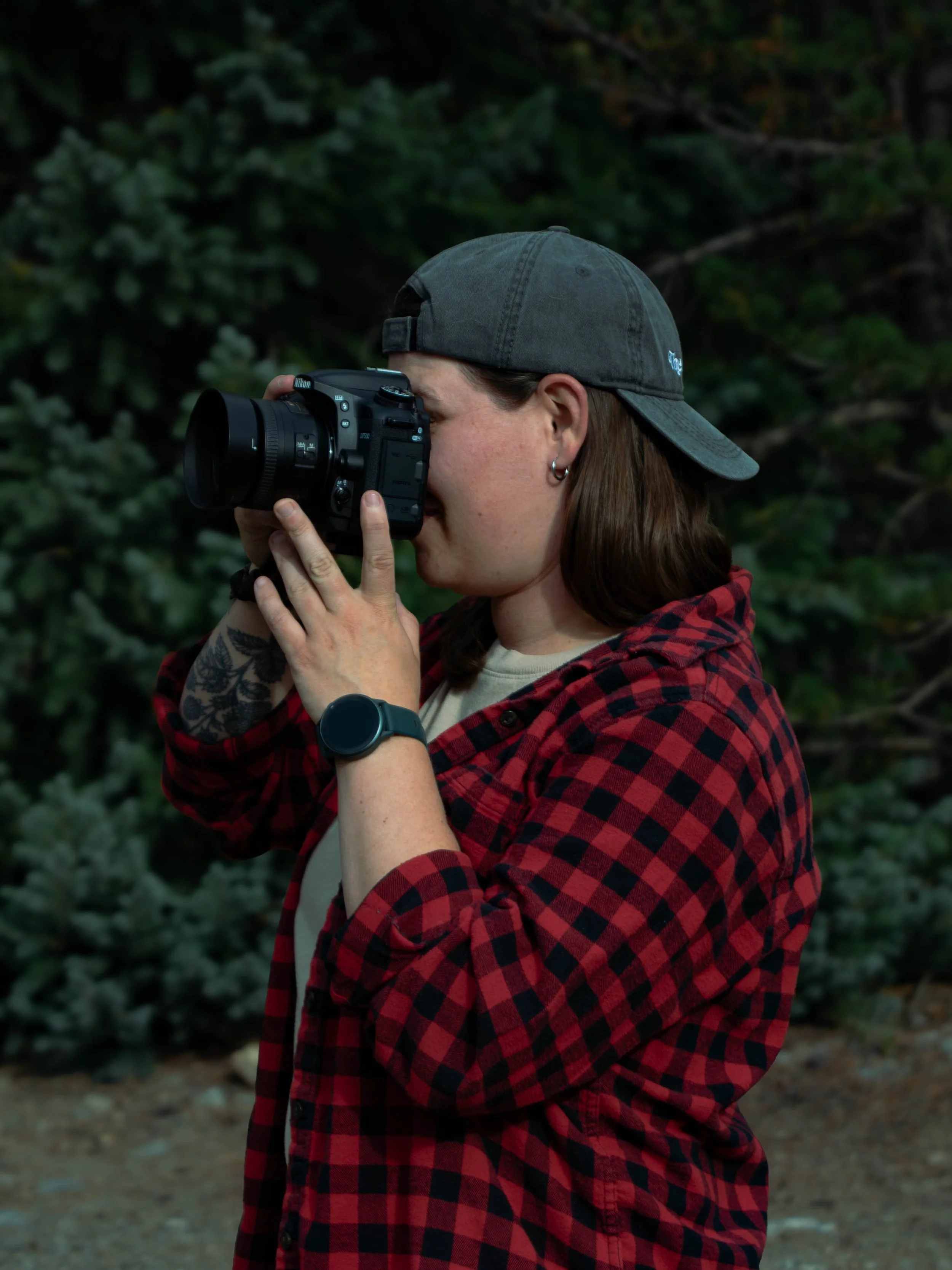 A person wearing a gray cap and red plaid shirt is taking photos with a black Nikon camera outdoors surrounded by green trees.