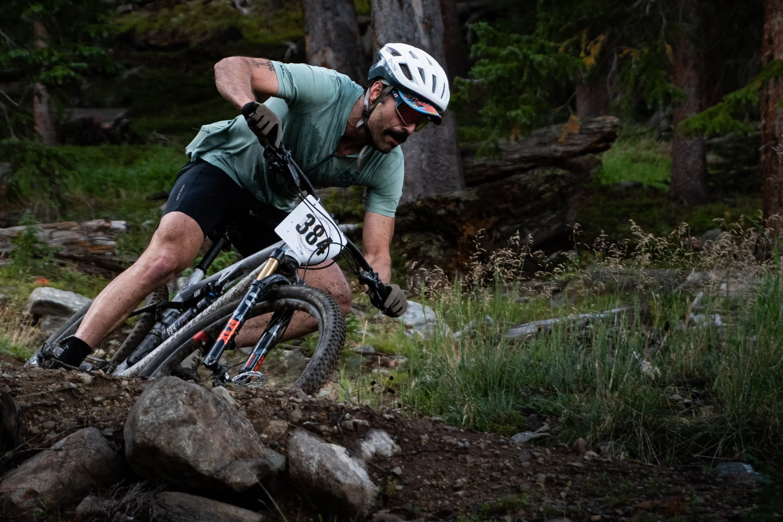 Man mountain biking downhill on a rocky trail in a forested area, wearing a helmet, sunglasses, and riding gear.