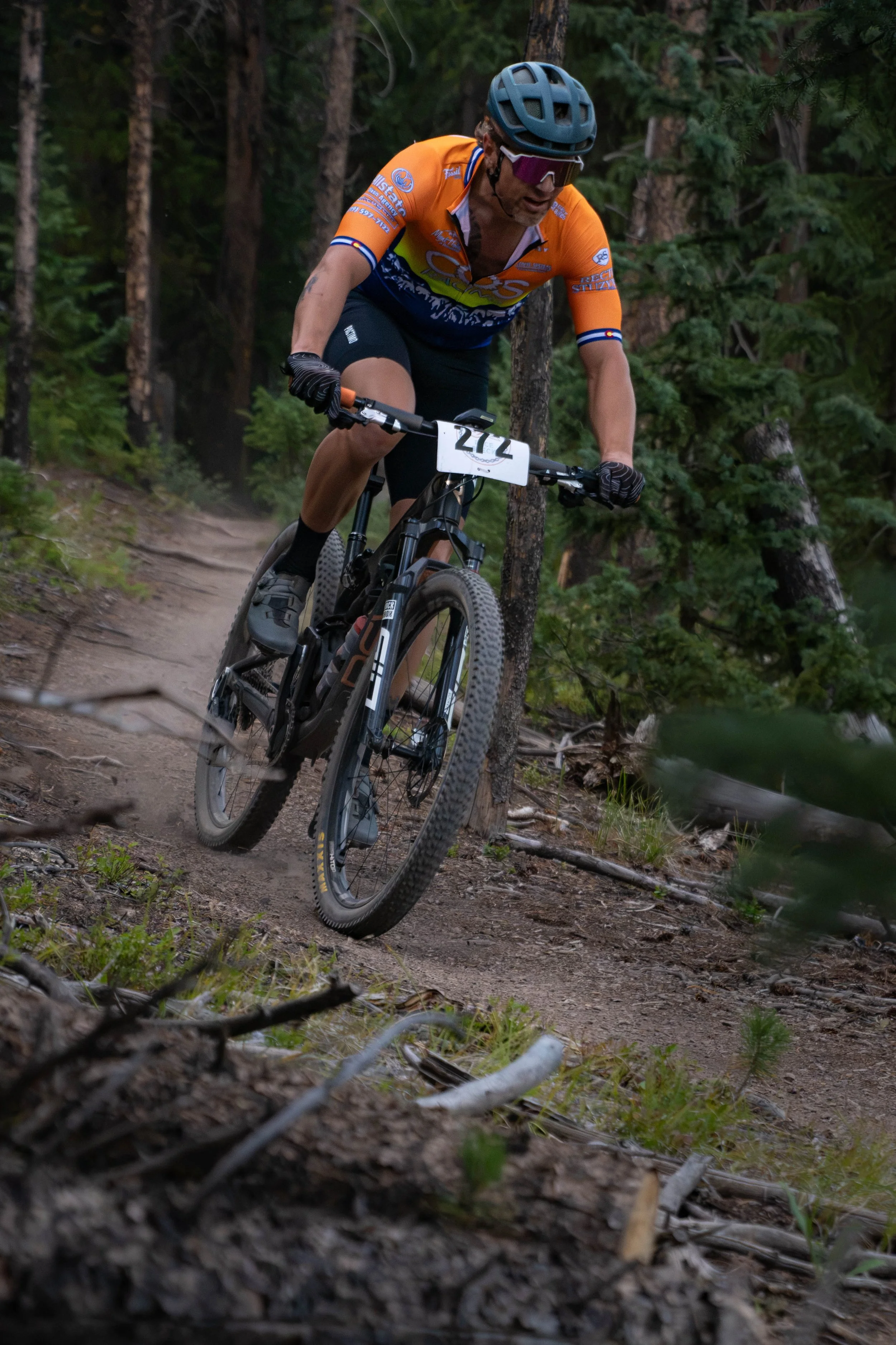 A mountain biker wearing an orange and black jersey, helmet, and sunglasses navigates a dirt trail through a forest with tall trees.