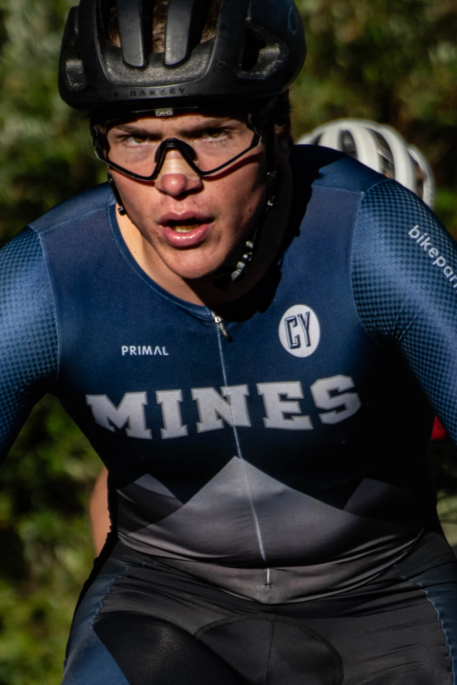 Close-up of a male cyclist wearing a black helmet and blue cycling jersey with the word 'MINES' visible, concentrating intensely while riding outdoors.